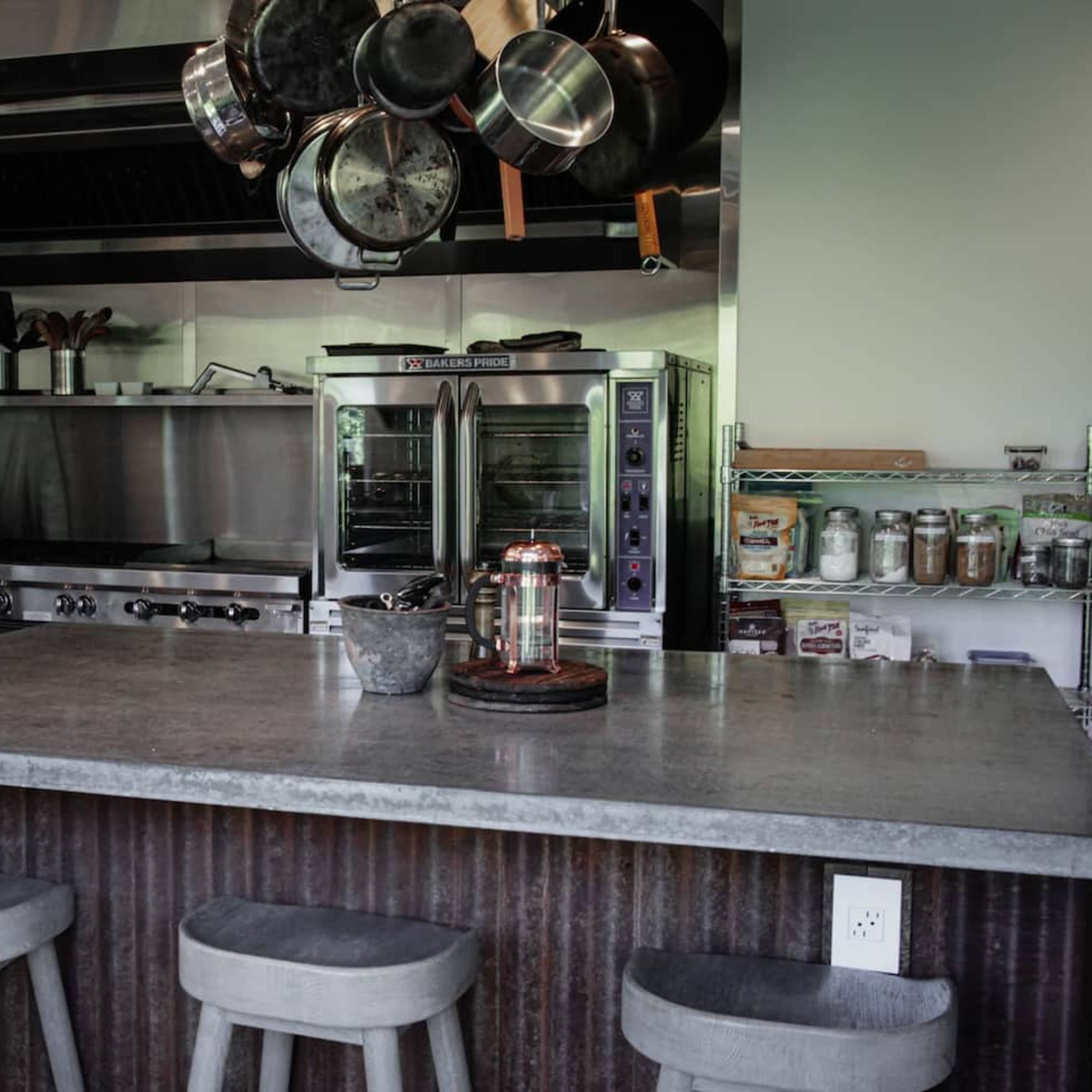 The kitchen features stainless steel appliances, a large concrete countertop, and a collection of hanging pots above an island with stool seating.