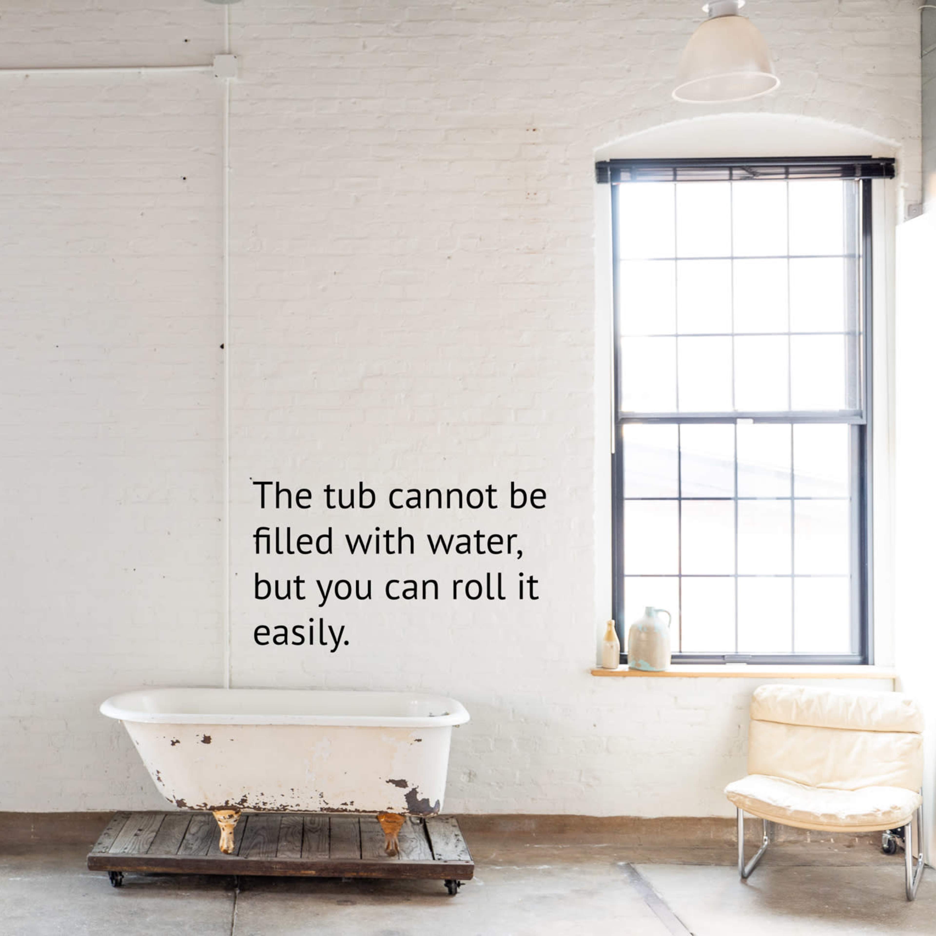 A minimalist room with large windows, a vintage freestanding bathtub on a wooden platform, a small desk with a chair, and a potted plant.