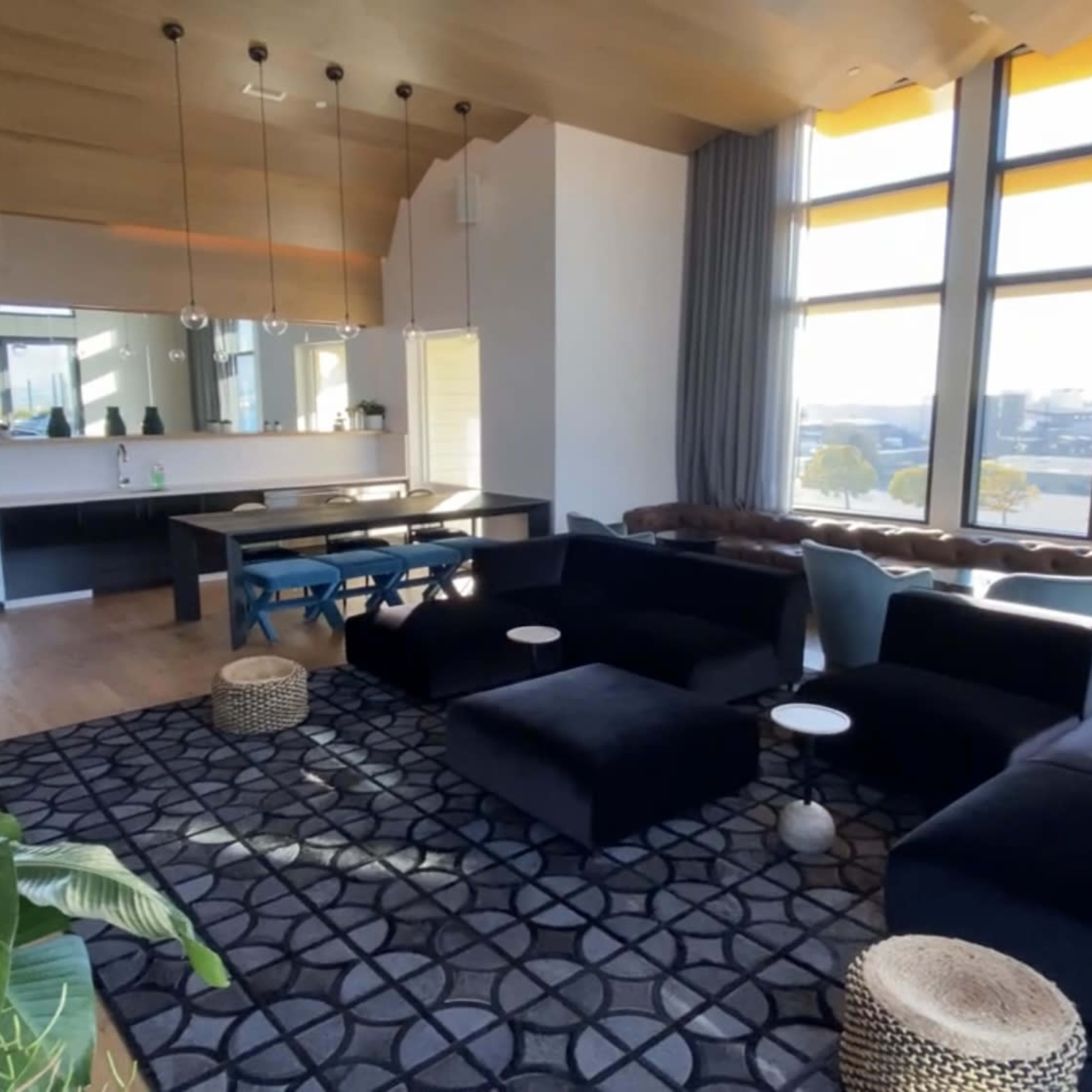 A modern lounge area featuring dark furniture, large windows with natural light, and a patterned rug.