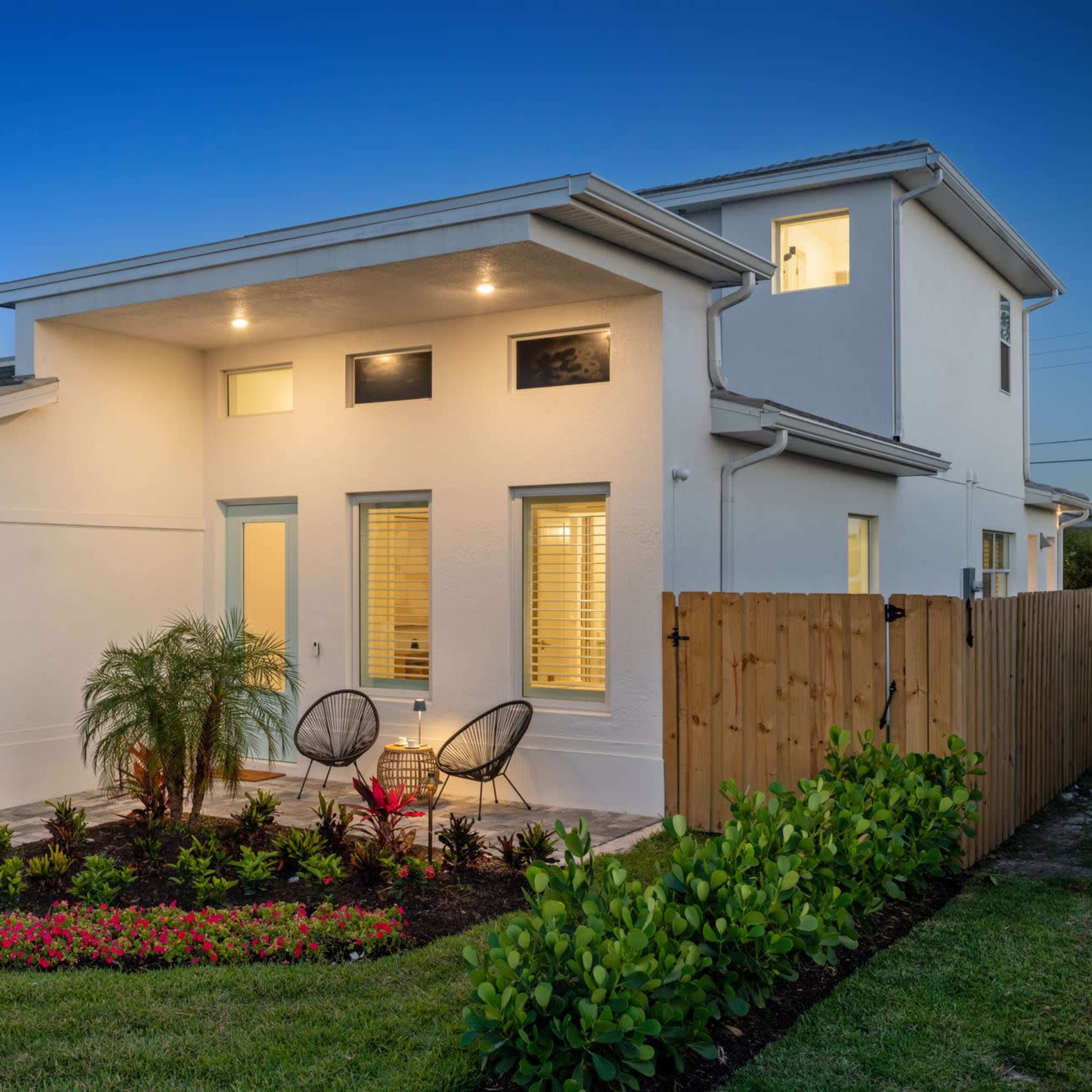 A modern, two-story white house with a well-lit exterior, a landscaped front yard, and a paved entrance featuring two chairs.