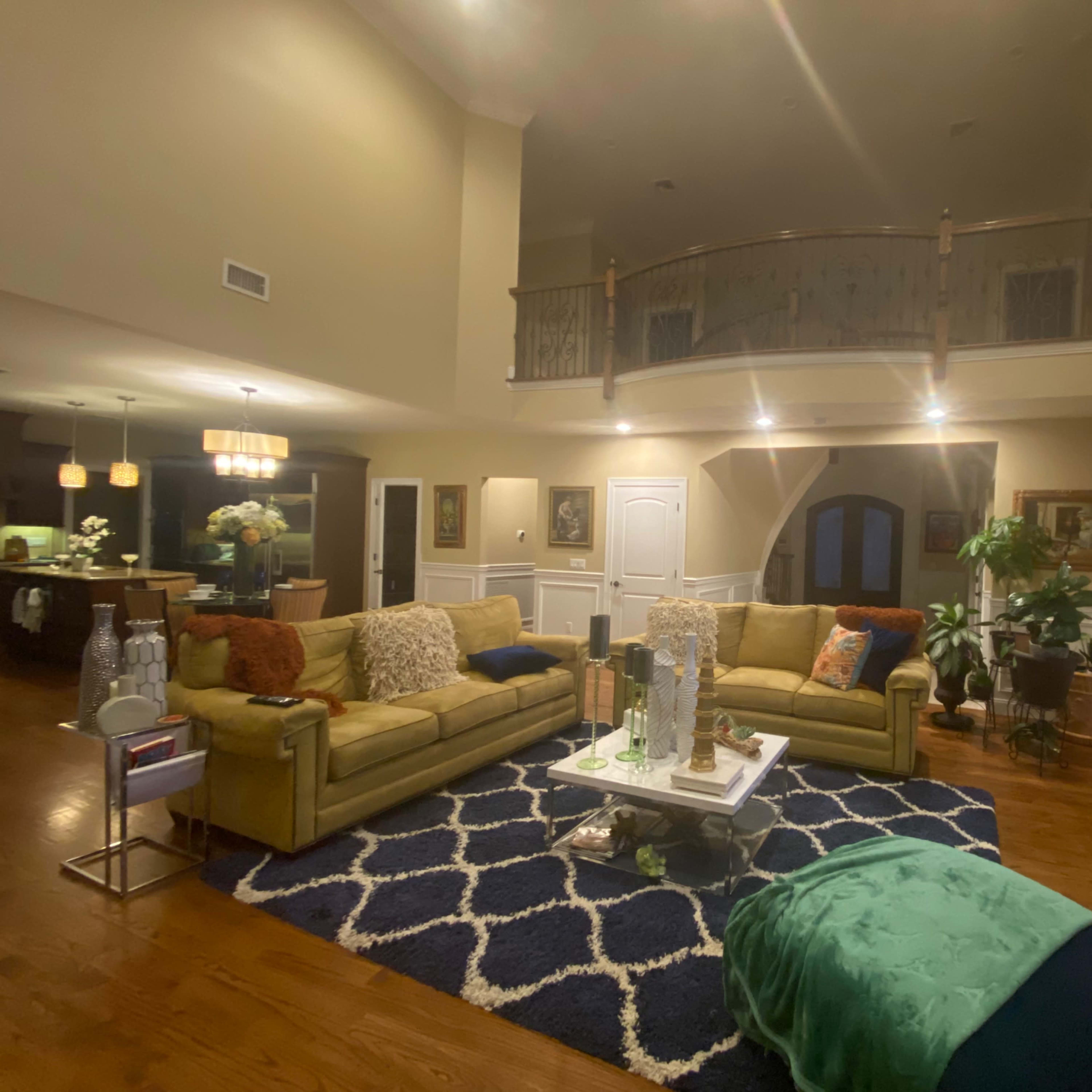 A spacious living room with two yellow sofas facing a coffee table, surrounded by artwork and decorative plants, and featuring a staircase leading to a balcony.