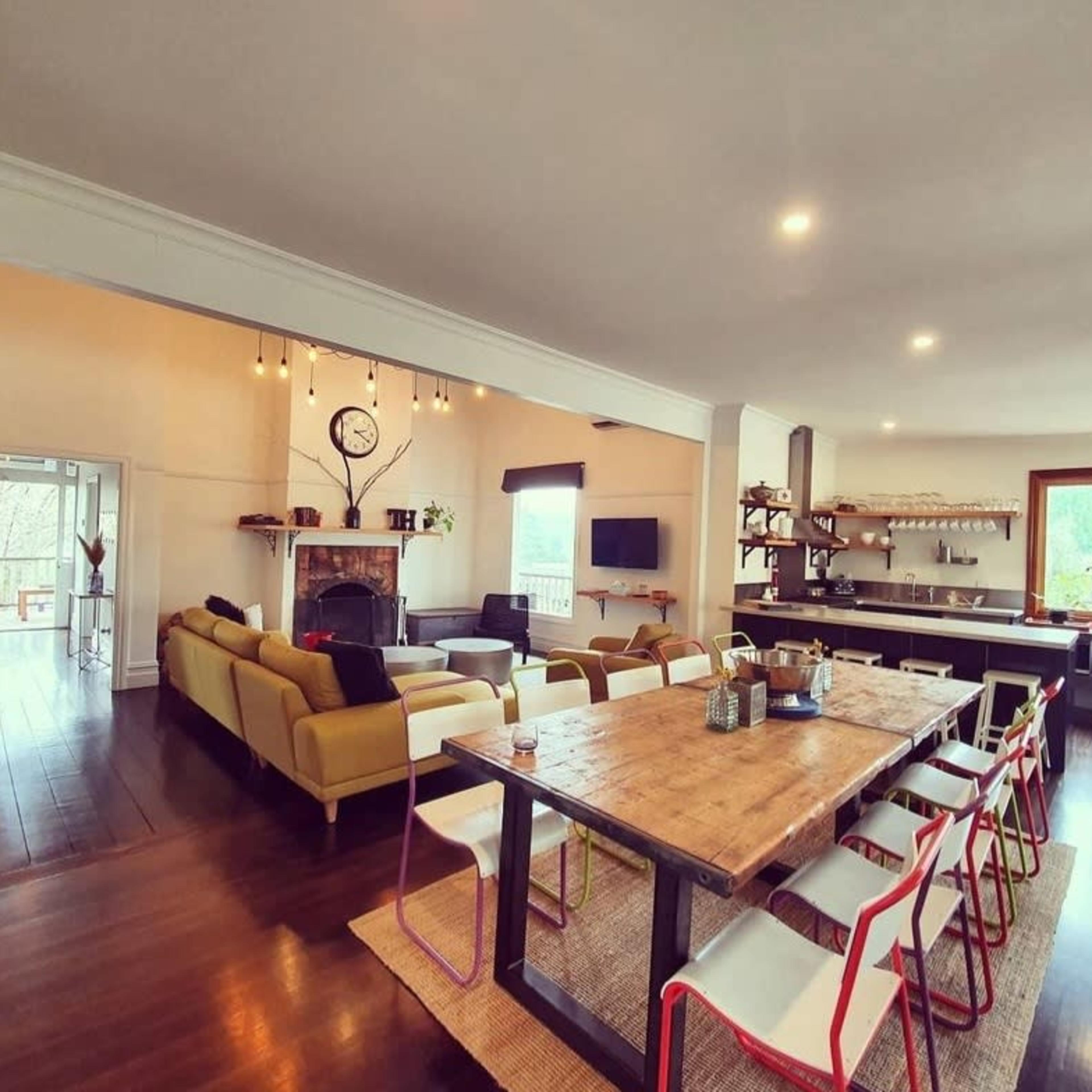 An open-concept living and dining area featuring a long wooden table with colorful chairs, a cozy seating arrangement, and a modern kitchen in the background.