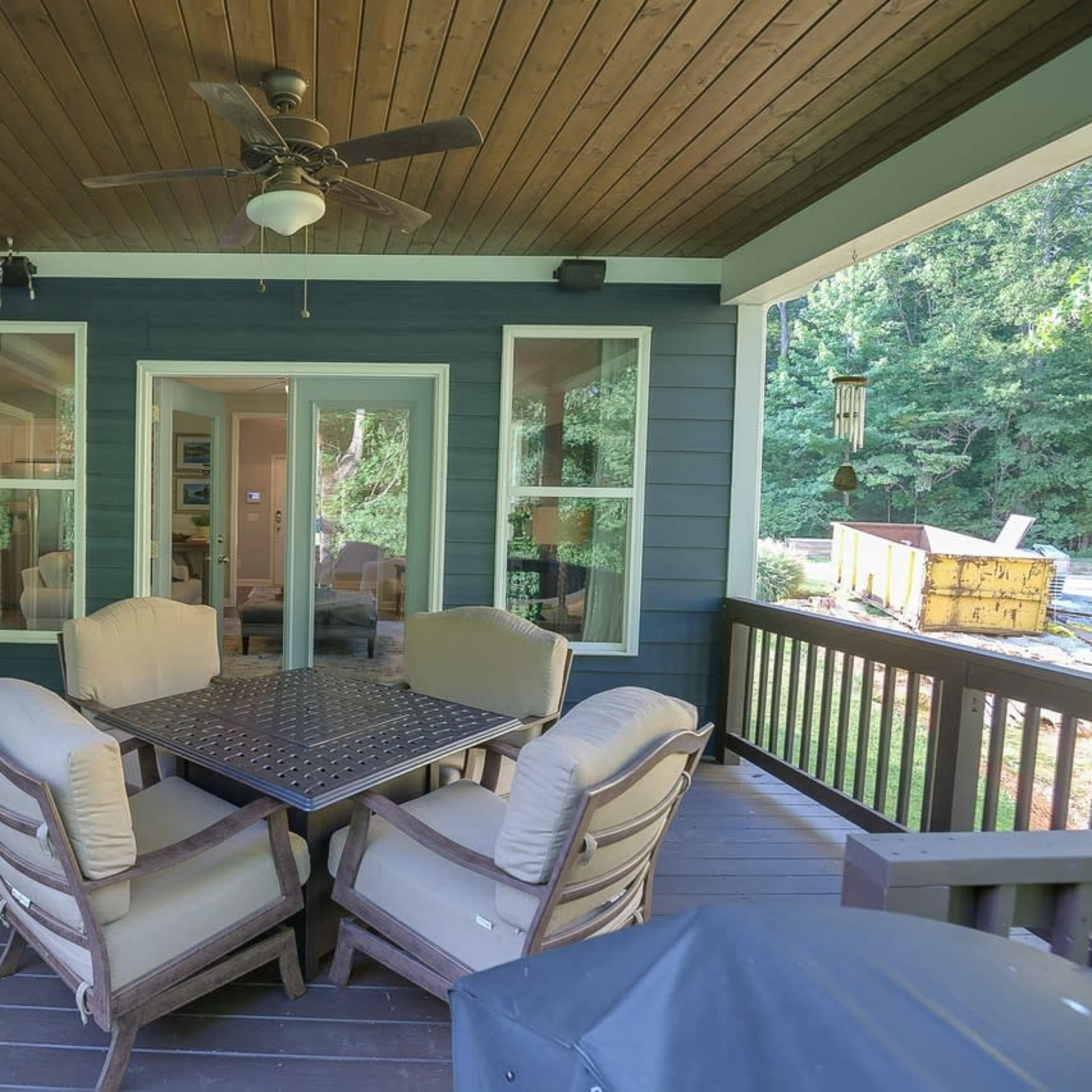 A covered porch with a table and four cushioned chairs, surrounded by windows and a view of greenery.