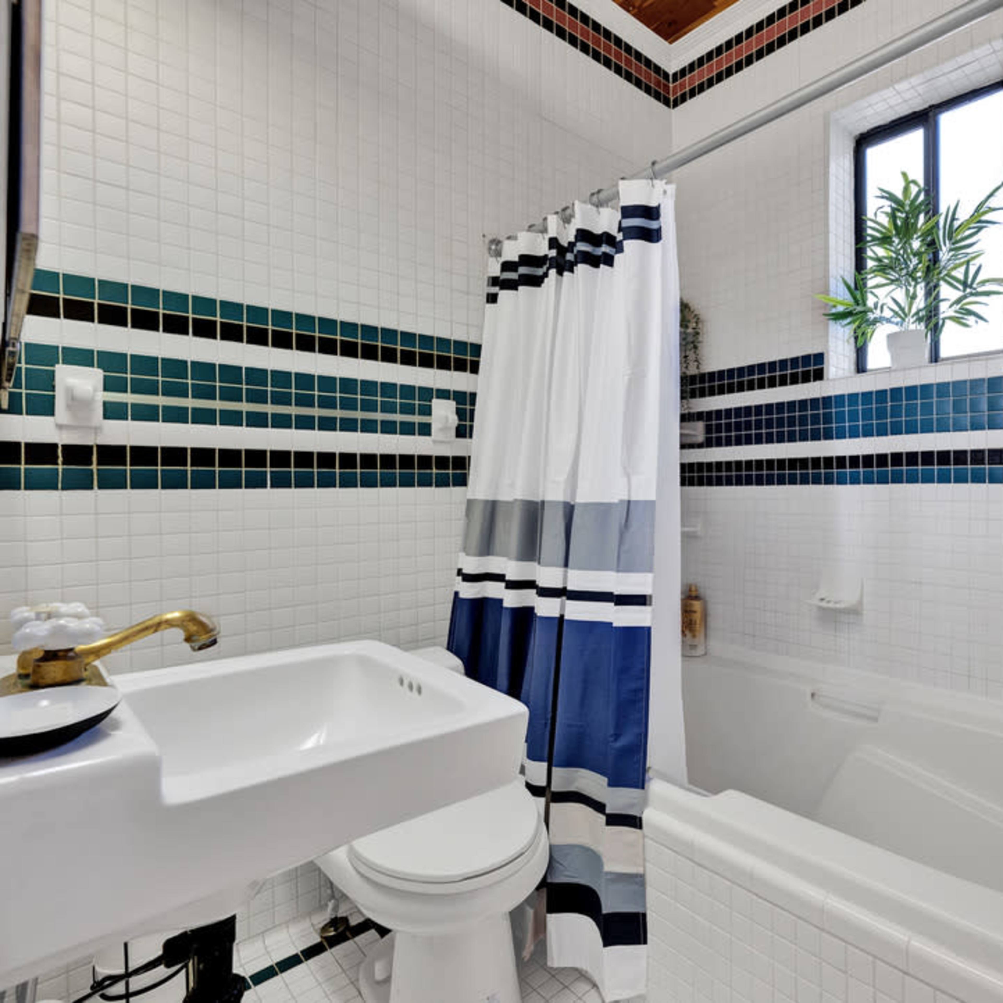 A tiled bathroom featuring a bathtub, a toilet, and a sink, with a striped shower curtain and a small plant by the window.