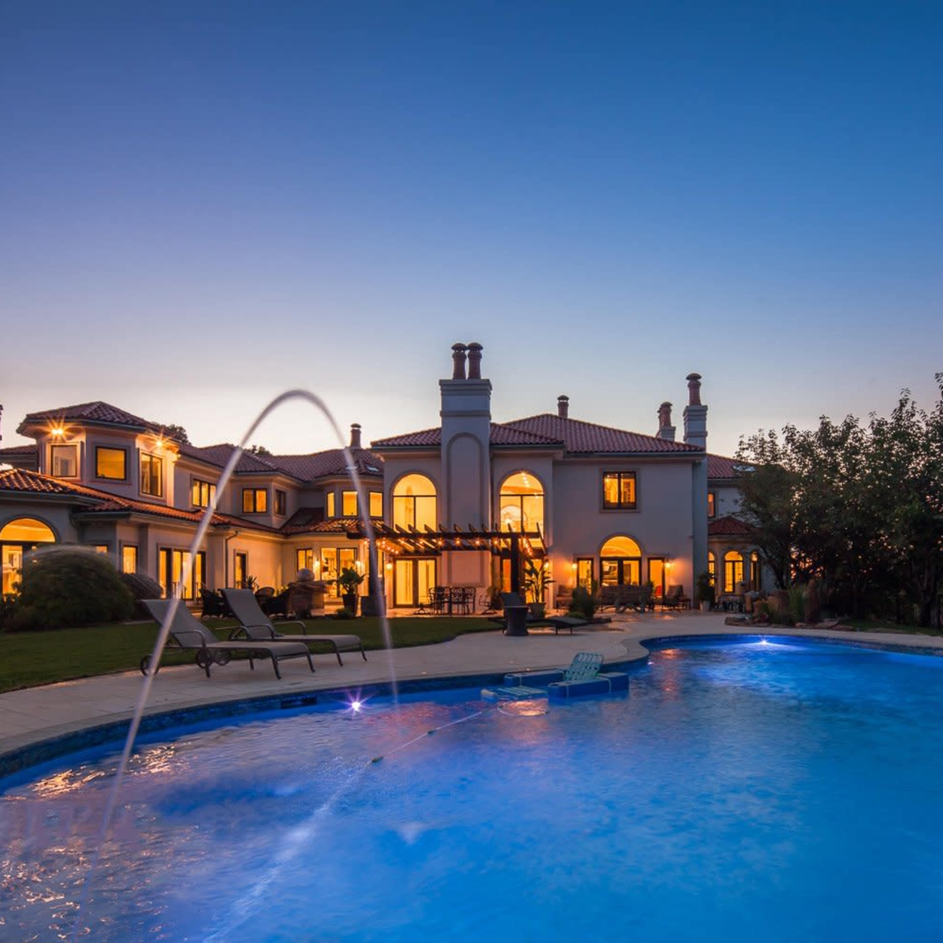 A large, elegant house with multiple windows and a balcony, illuminated at dusk, alongside a blue swimming pool featuring a fountain.