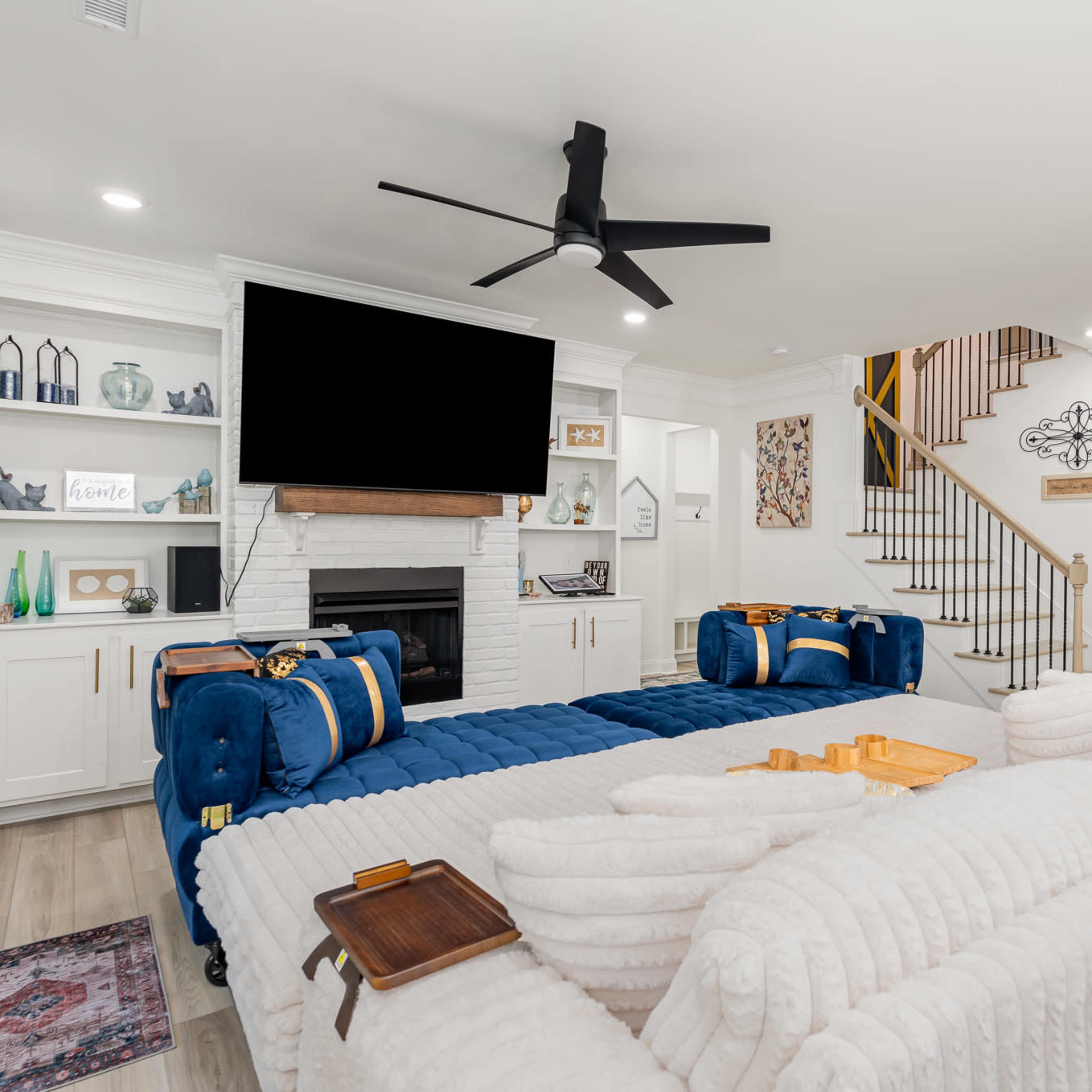 A modern living room features a central seating area with a white sectional sofa and blue accent chairs, a large flat-screen TV above a fireplace, and a staircase in the background.