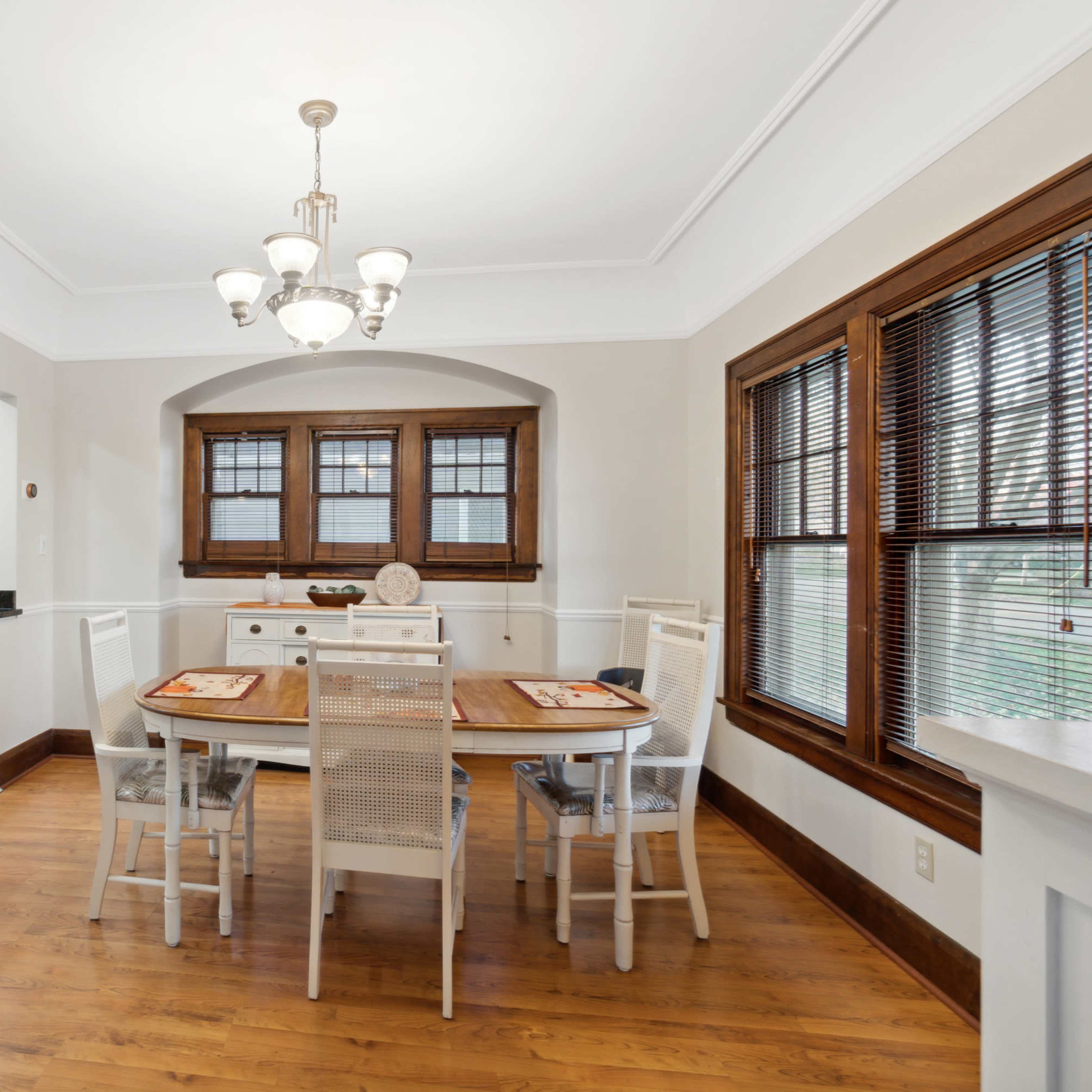 A dining area features a wooden table surrounded by white chairs, with large windows and a chandelier overhead.