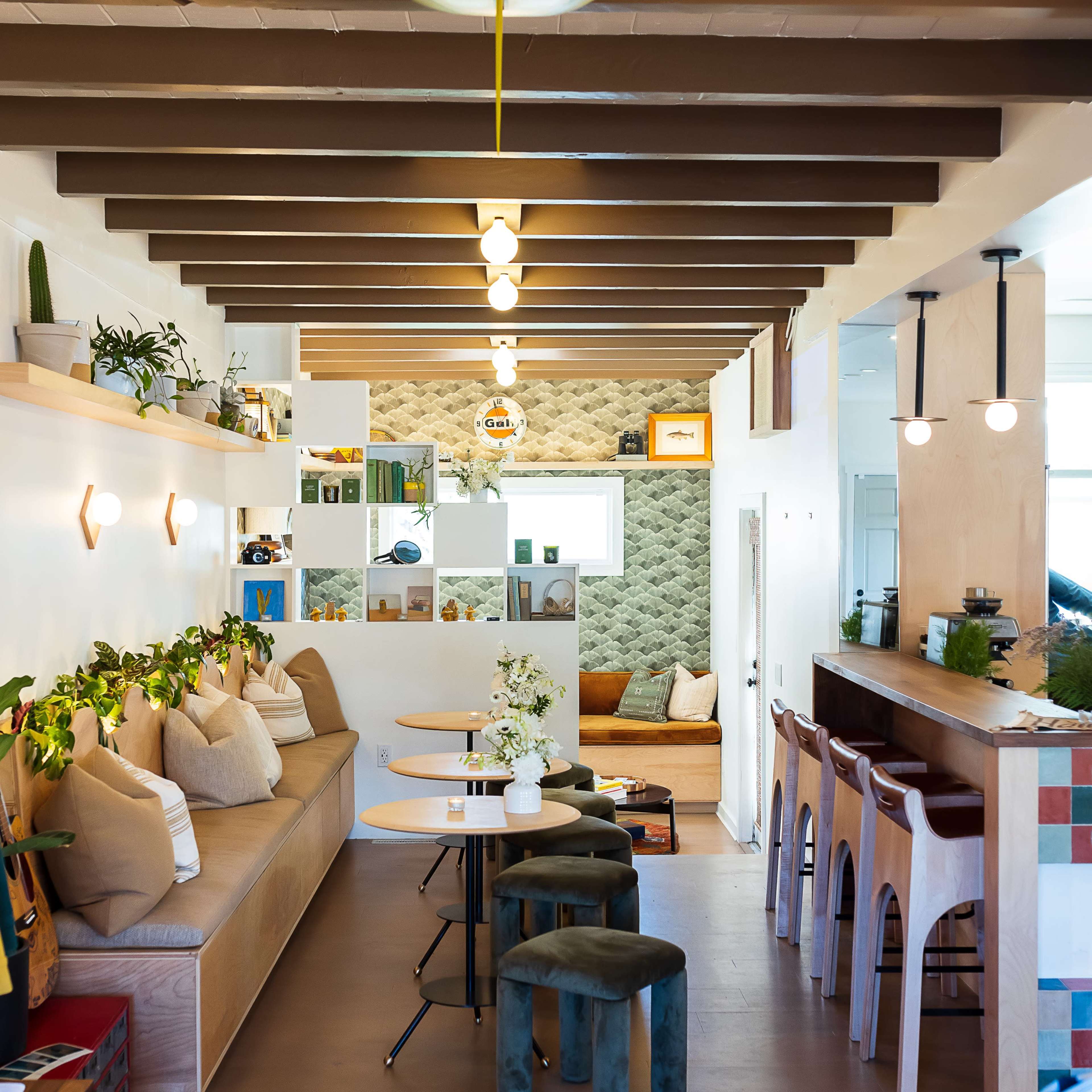 A modern café interior with wooden beams, green accents, seating areas with cushions, and a surfboard hanging from the ceiling.