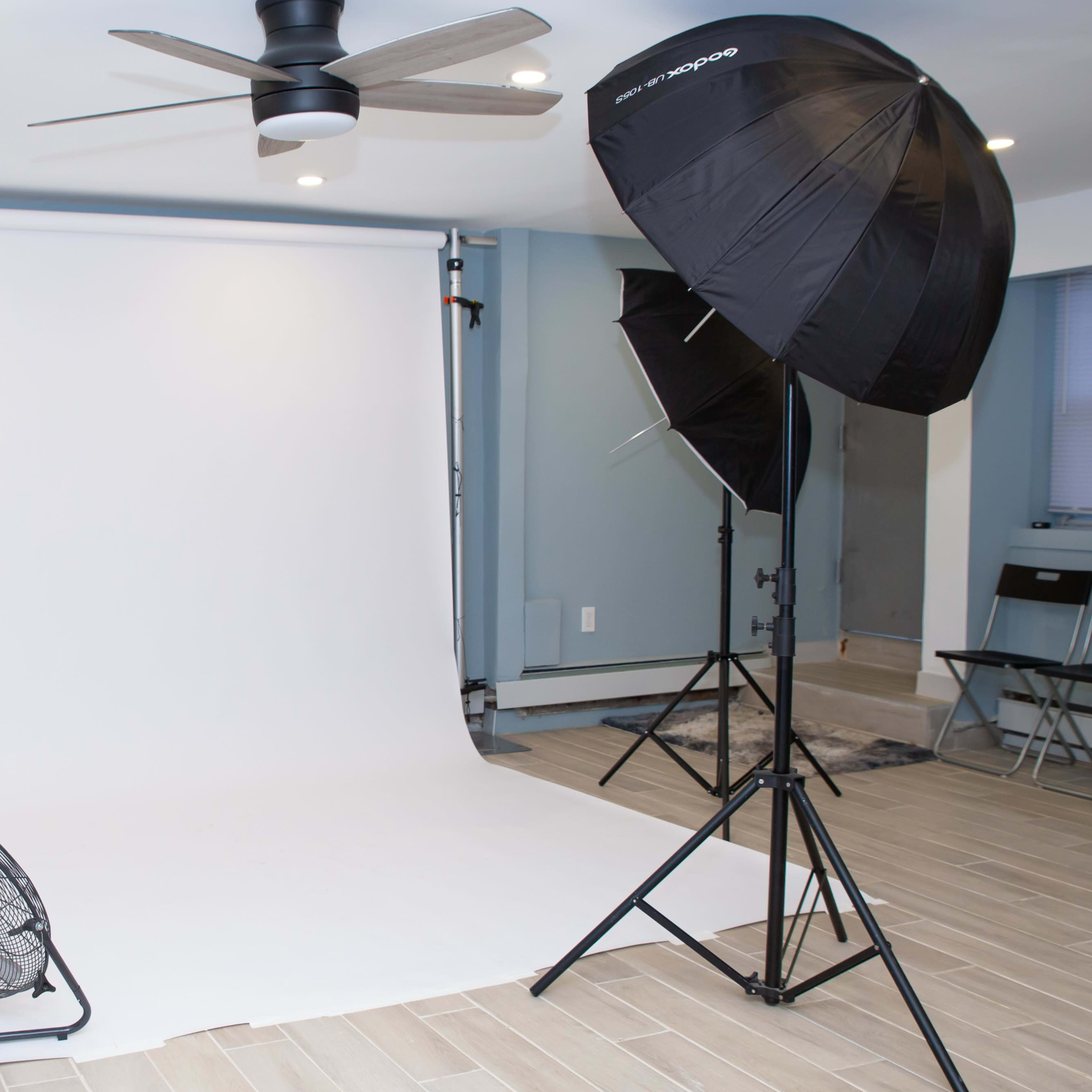 A photo studio setup with a white backdrop, two softbox lights, and a fan positioned on the floor.