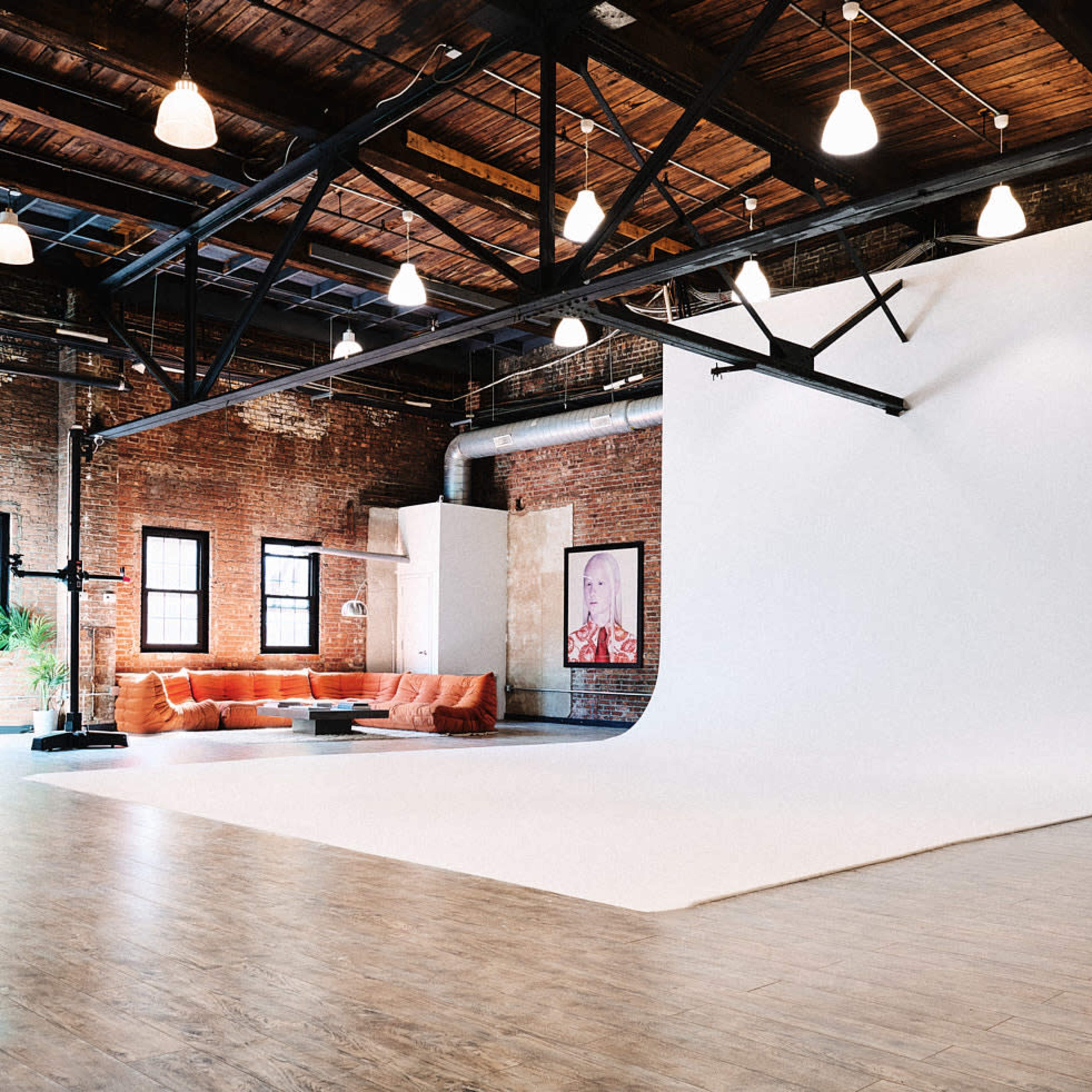 A spacious studio with exposed brick walls, a white backdrop, and a lounge area featuring orange couches.