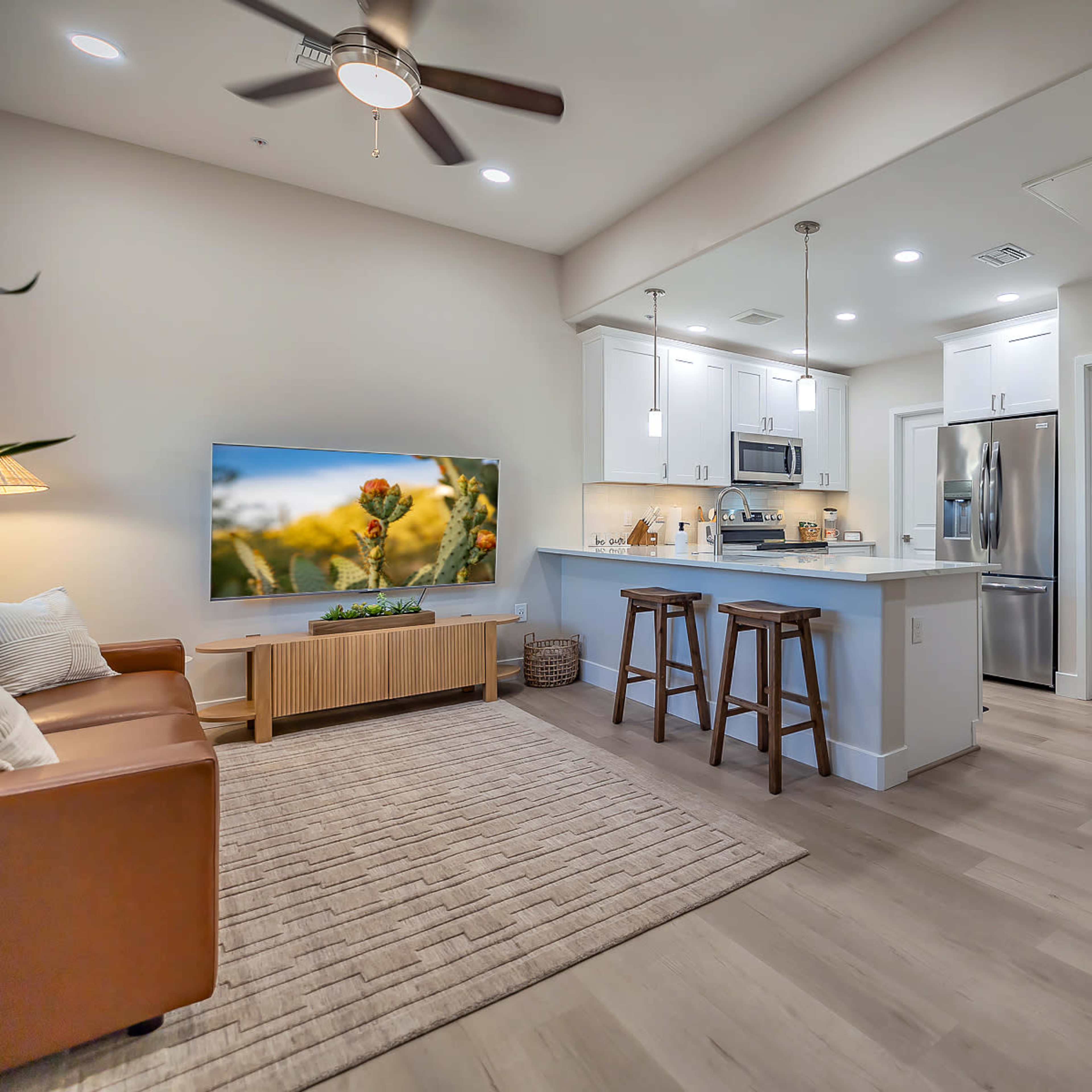 A modern living area with a kitchen, featuring a brown leather sofa, a circular dining table, and a wall-mounted television.