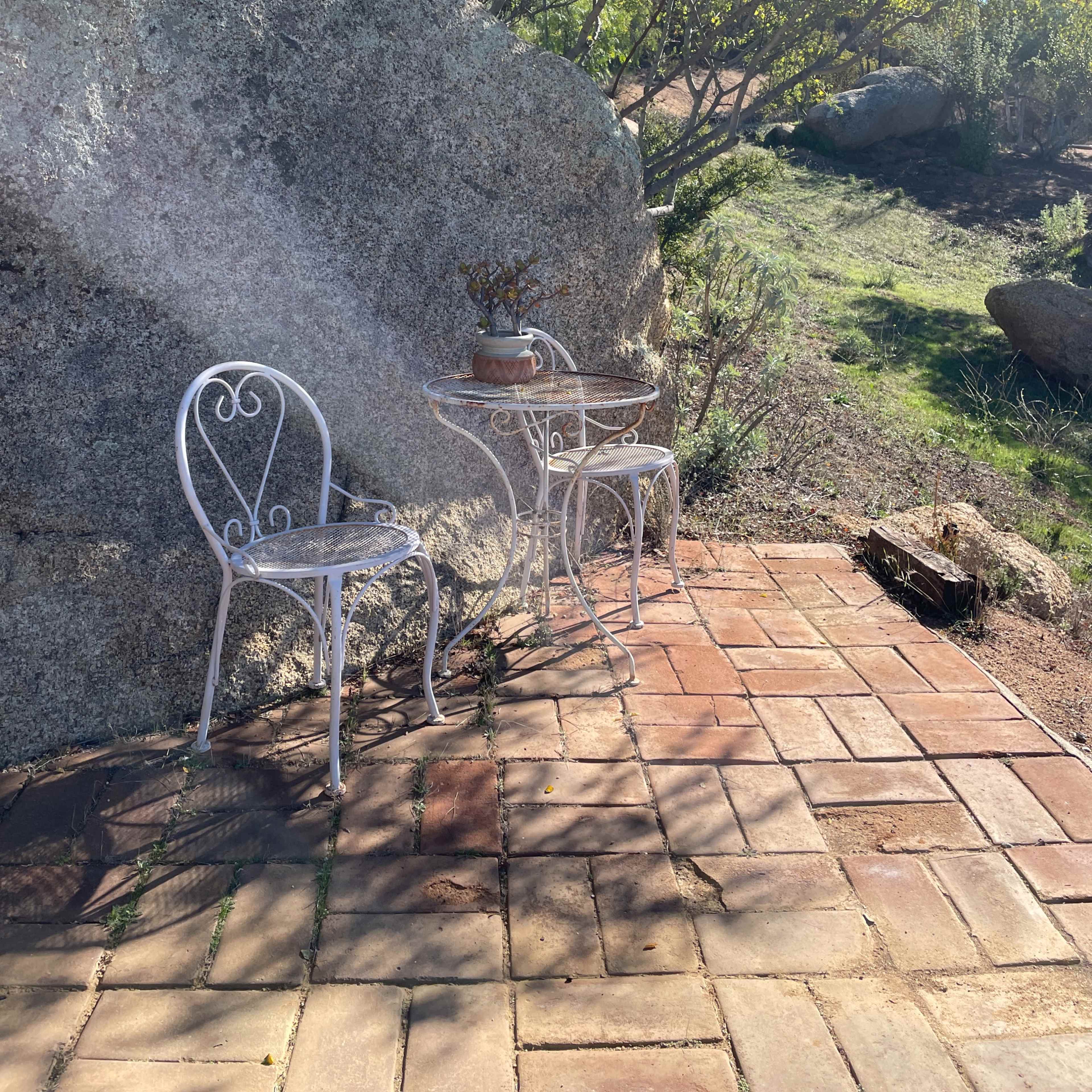 A small outdoor seating area with two white metal chairs and a table is positioned next to a large rock and surrounded by greenery.