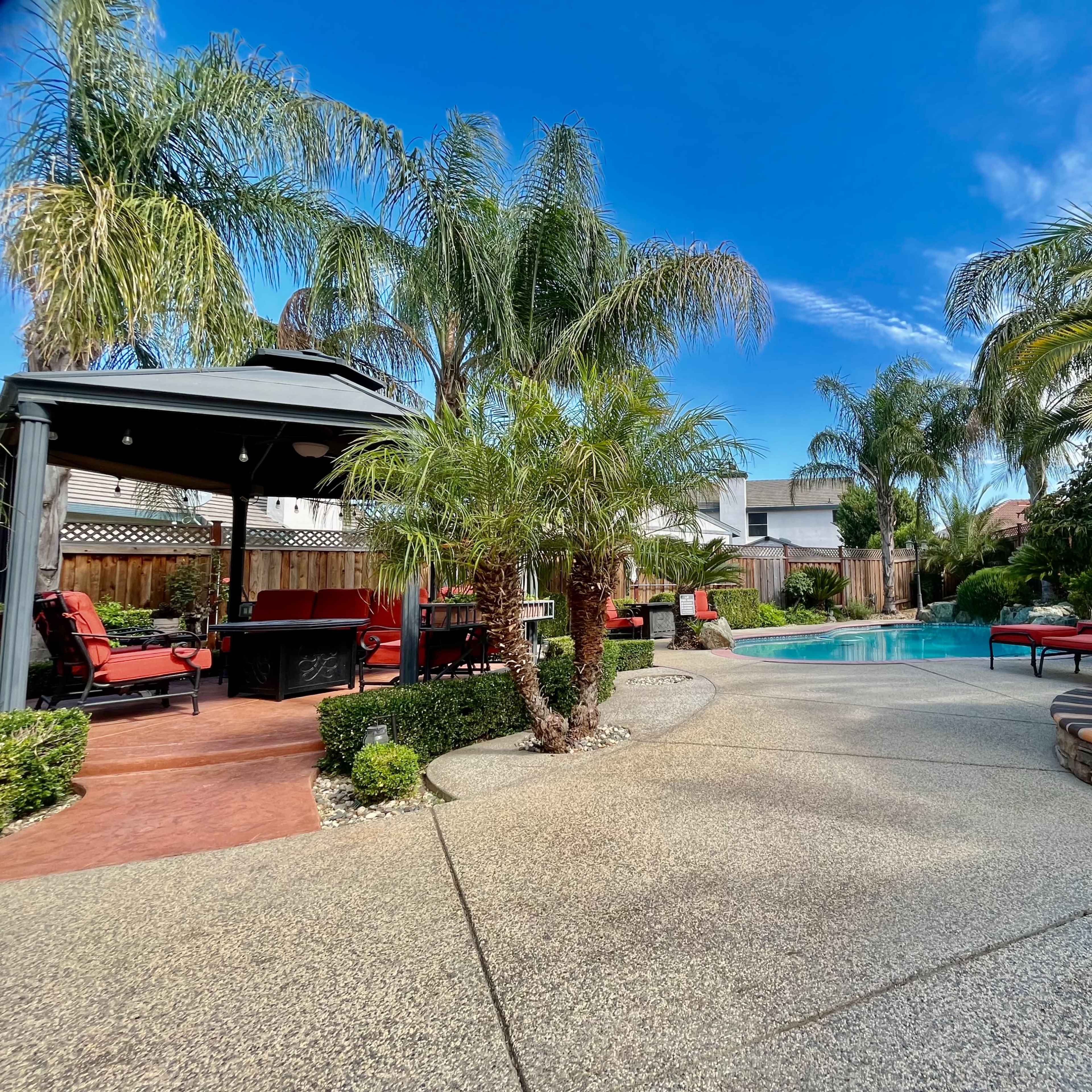 A backyard oasis featuring a swimming pool surrounded by lounge chairs, palm trees, and a gazebo.