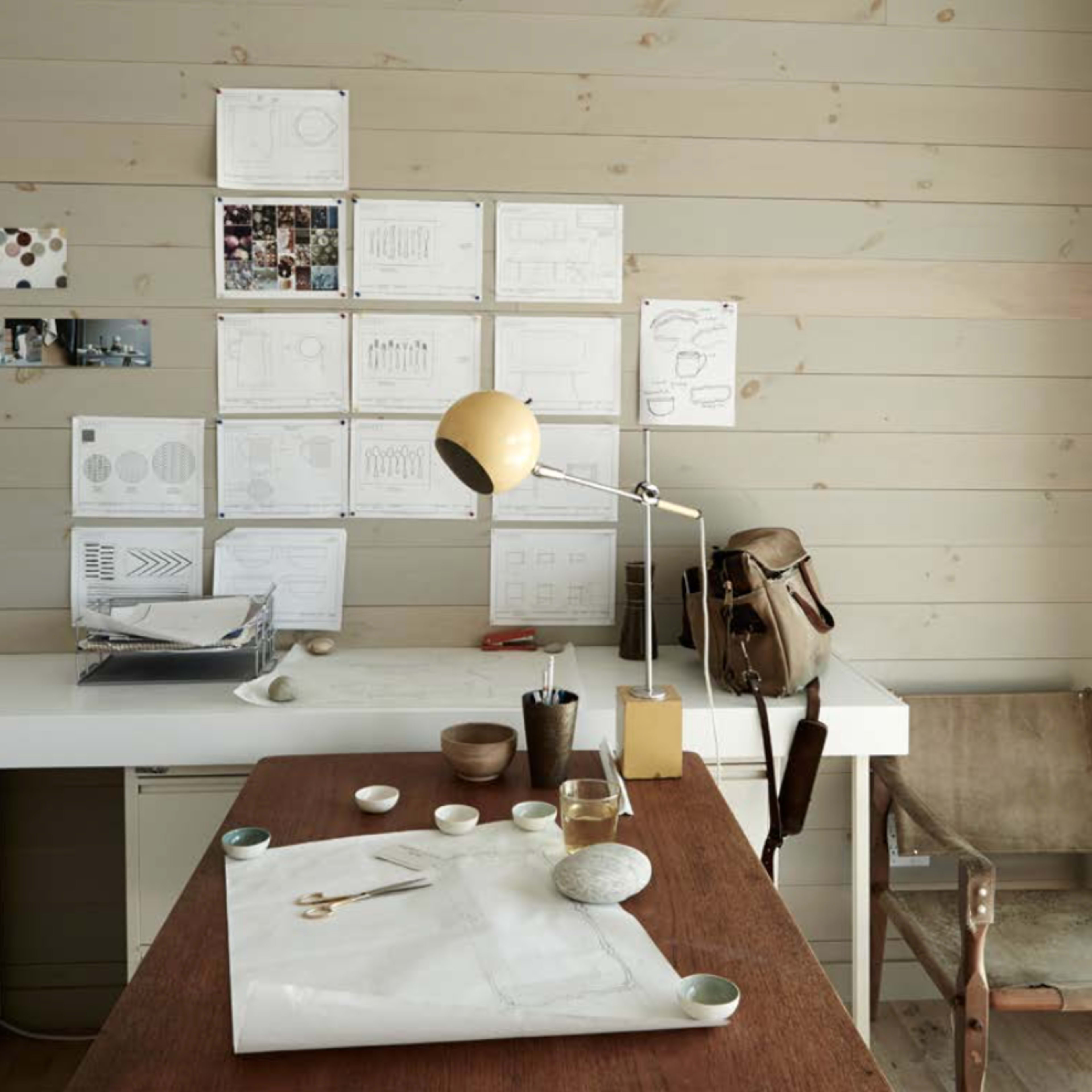 A workspace features a white table with design sketches pinned on a light wood wall, a desk lamp, and various small items neatly arranged.