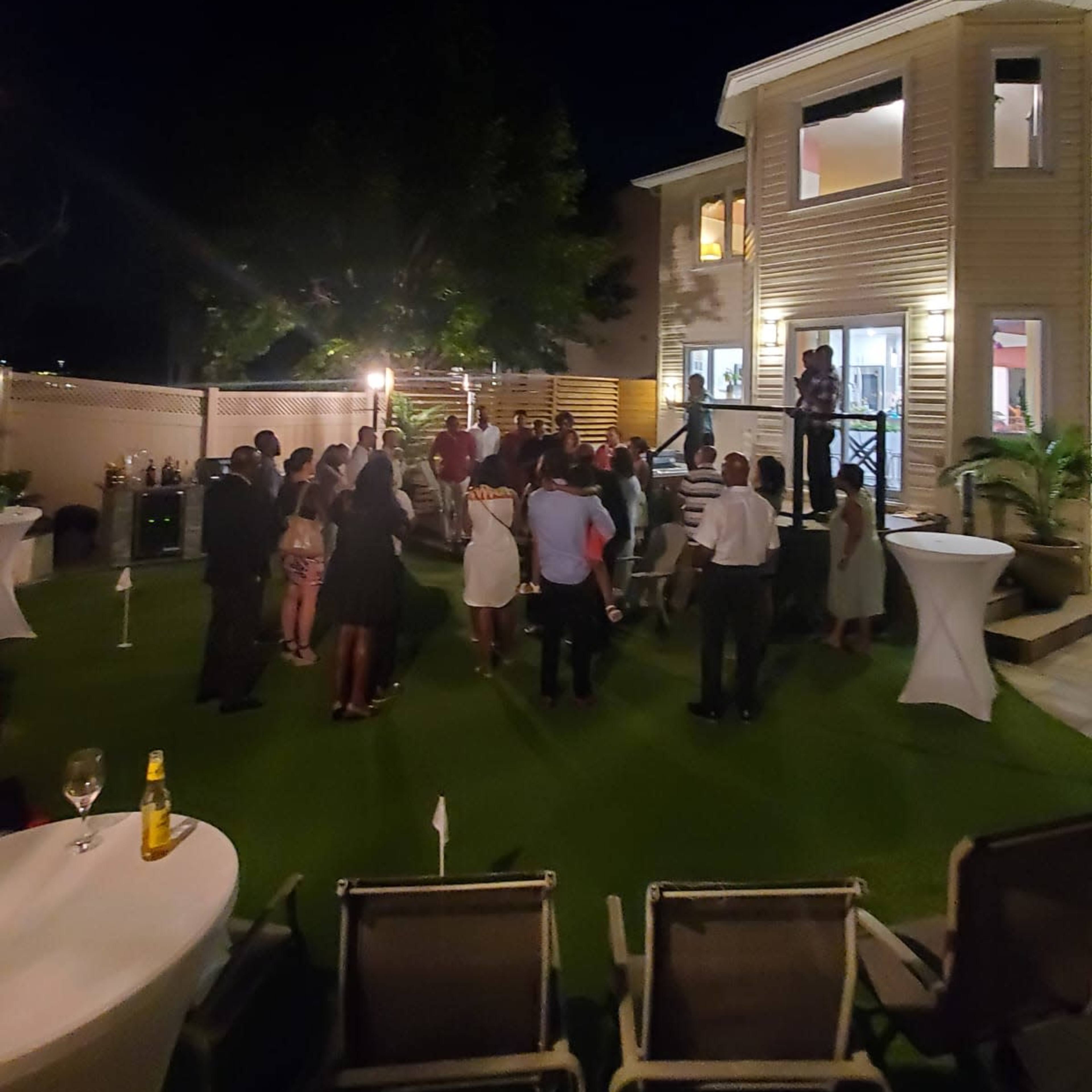 A group of people gathers on a well-lit patio at night, with a house in the background and tables set up for a social event.