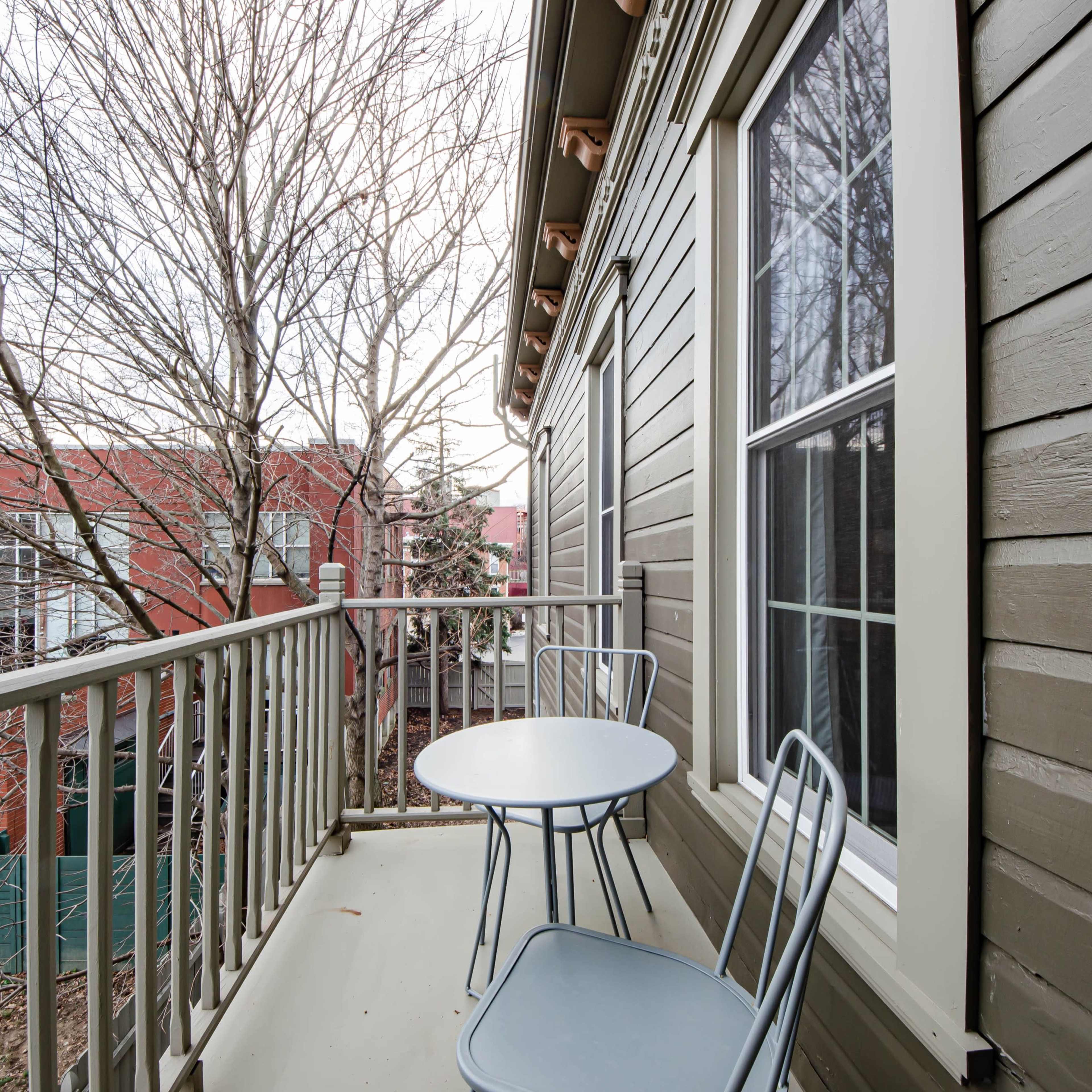 A small balcony with two metal chairs and a round table, surrounded by bare trees and a view of a red brick building.