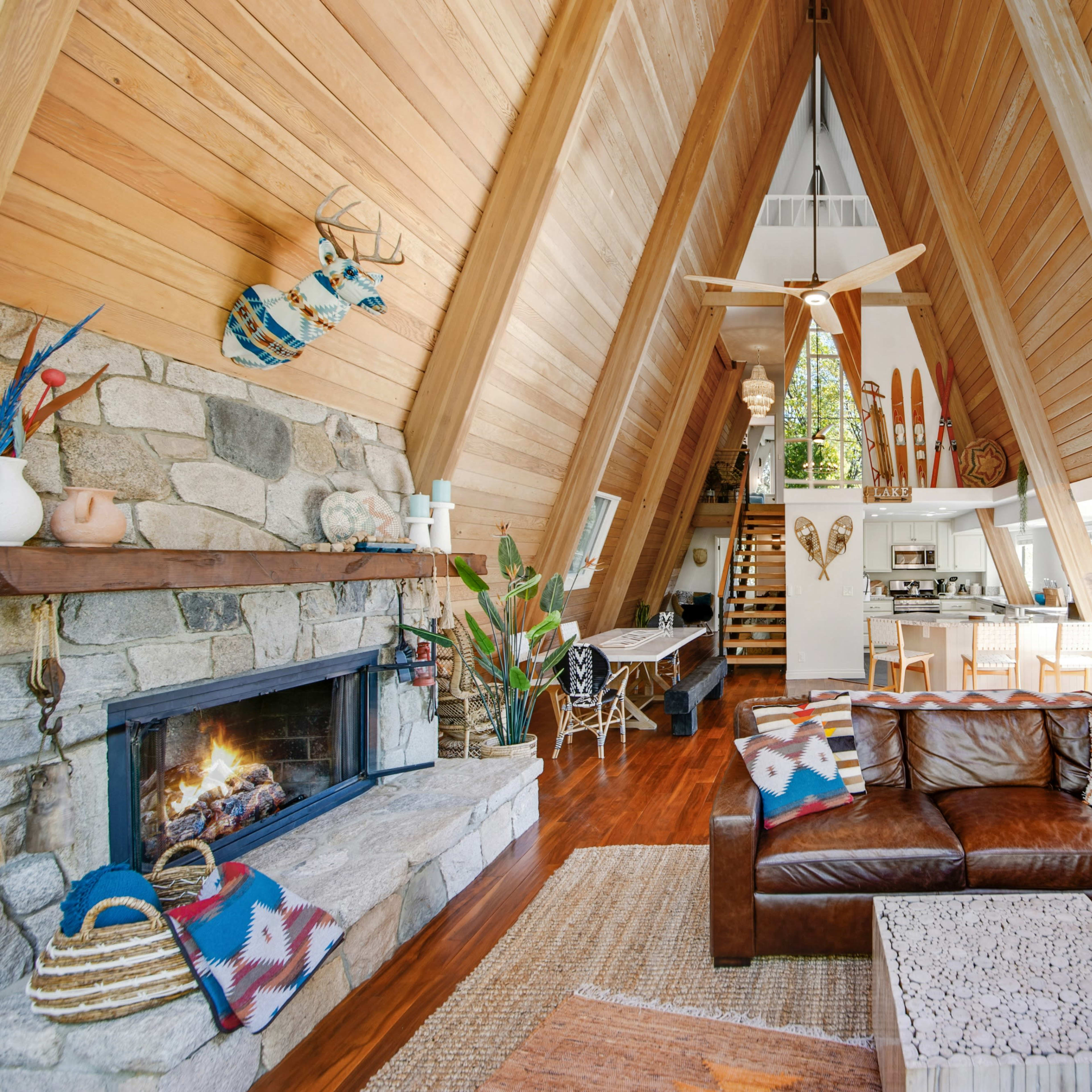A modern A-frame cabin interior with a wooden framework, a stone fireplace, and a spacious living area featuring a leather sectional sofa and a view of the outdoors.