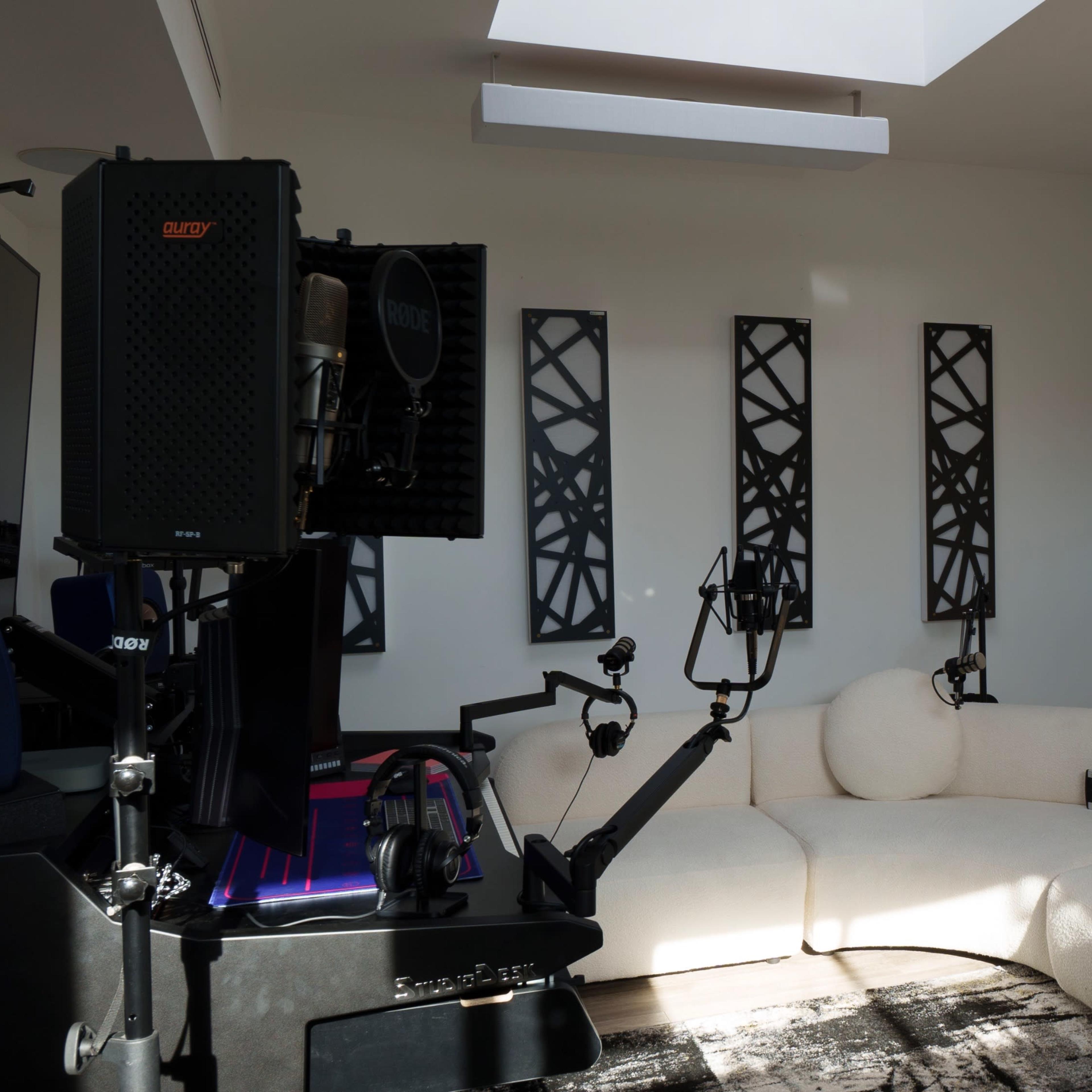 A modern room with a curved white sofa, three microphones mounted on stands, and large windows with partially opened blinds.