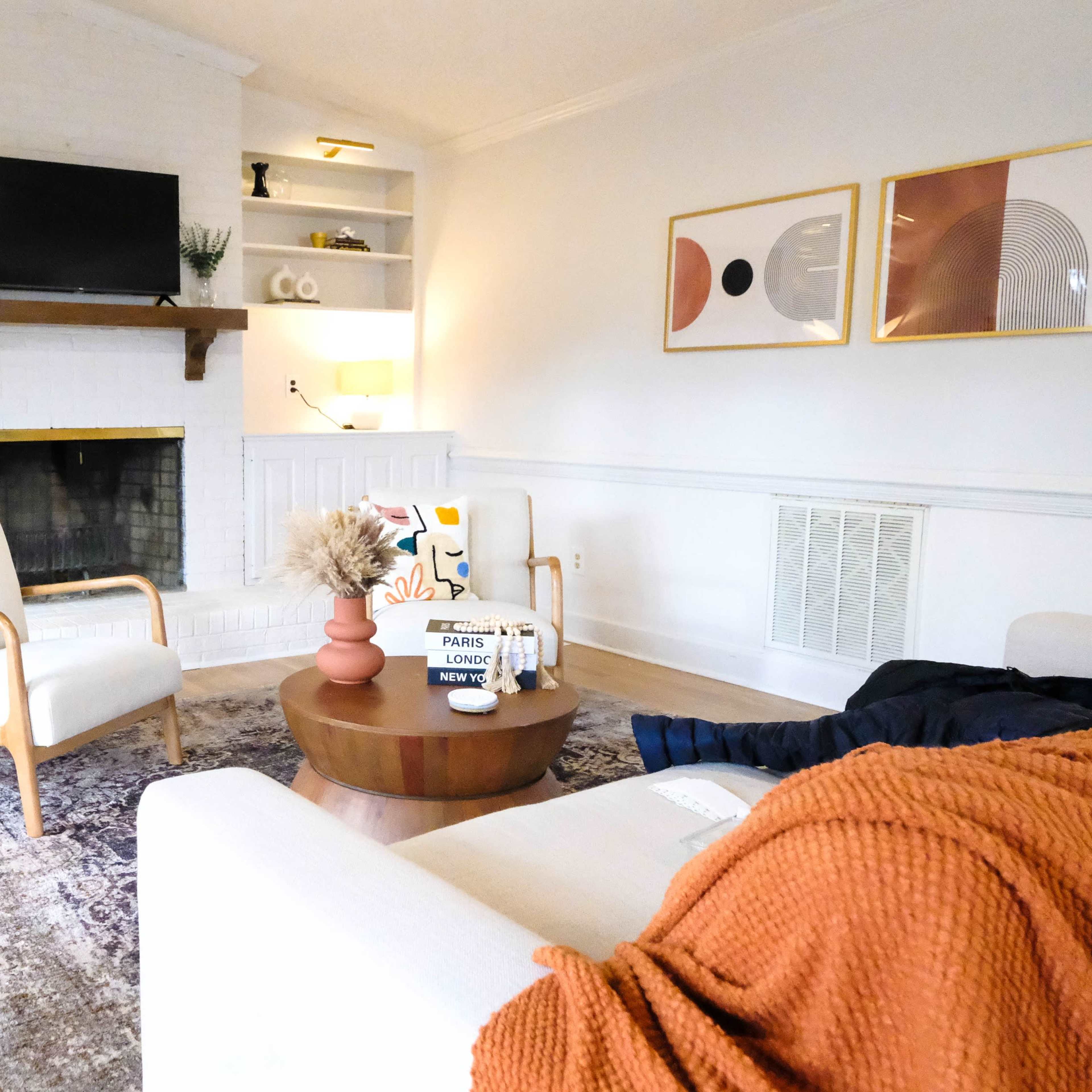 A bright living room featuring two armchairs, a coffee table, a fireplace, and a large TV mounted on the wall.
