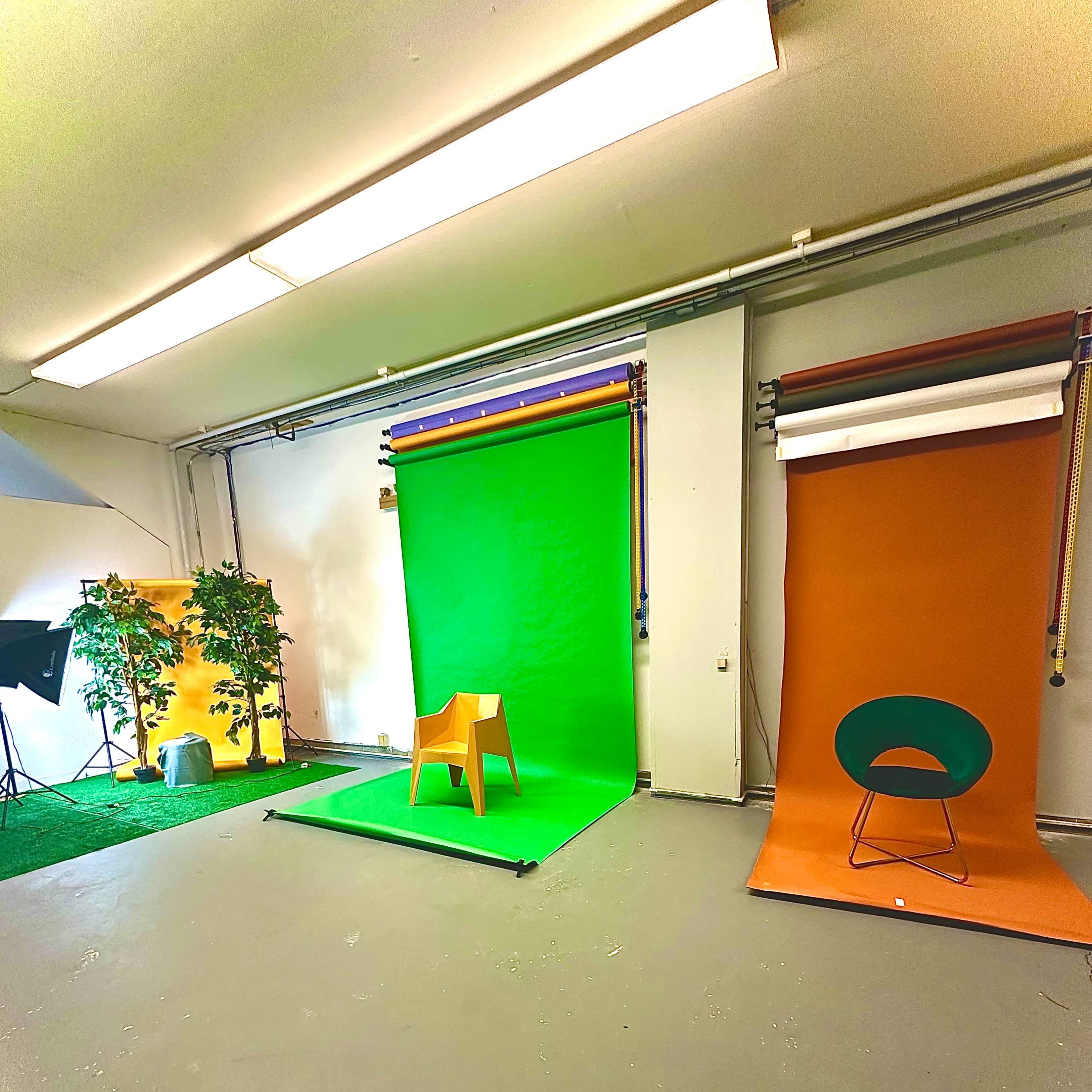 A photography studio with three colored backdrops: green, orange, and another behind a chair, alongside a potted tree and lighting equipment.