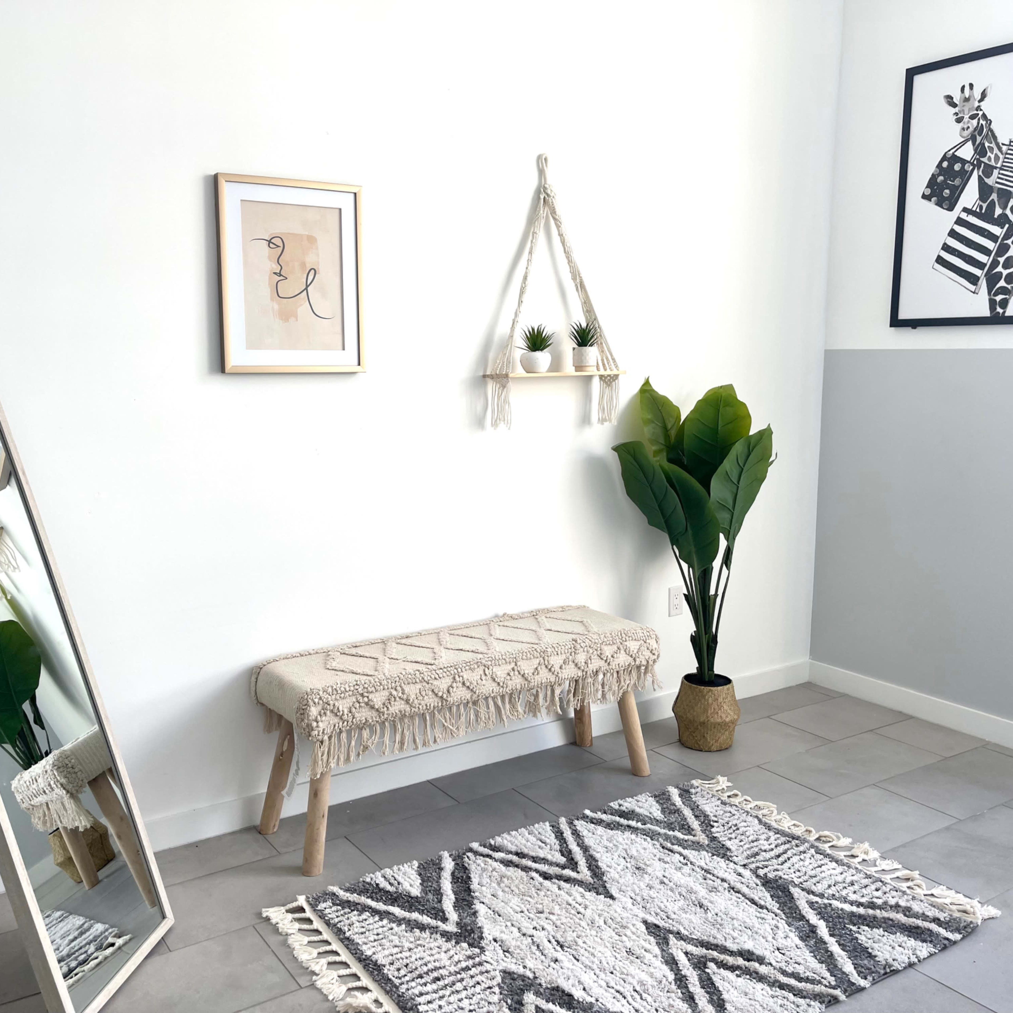 A minimalist room featuring a mirror, a bench with decorative fringe, a shelf with small plants, a plant in a pot, and two framed artworks on the walls.
