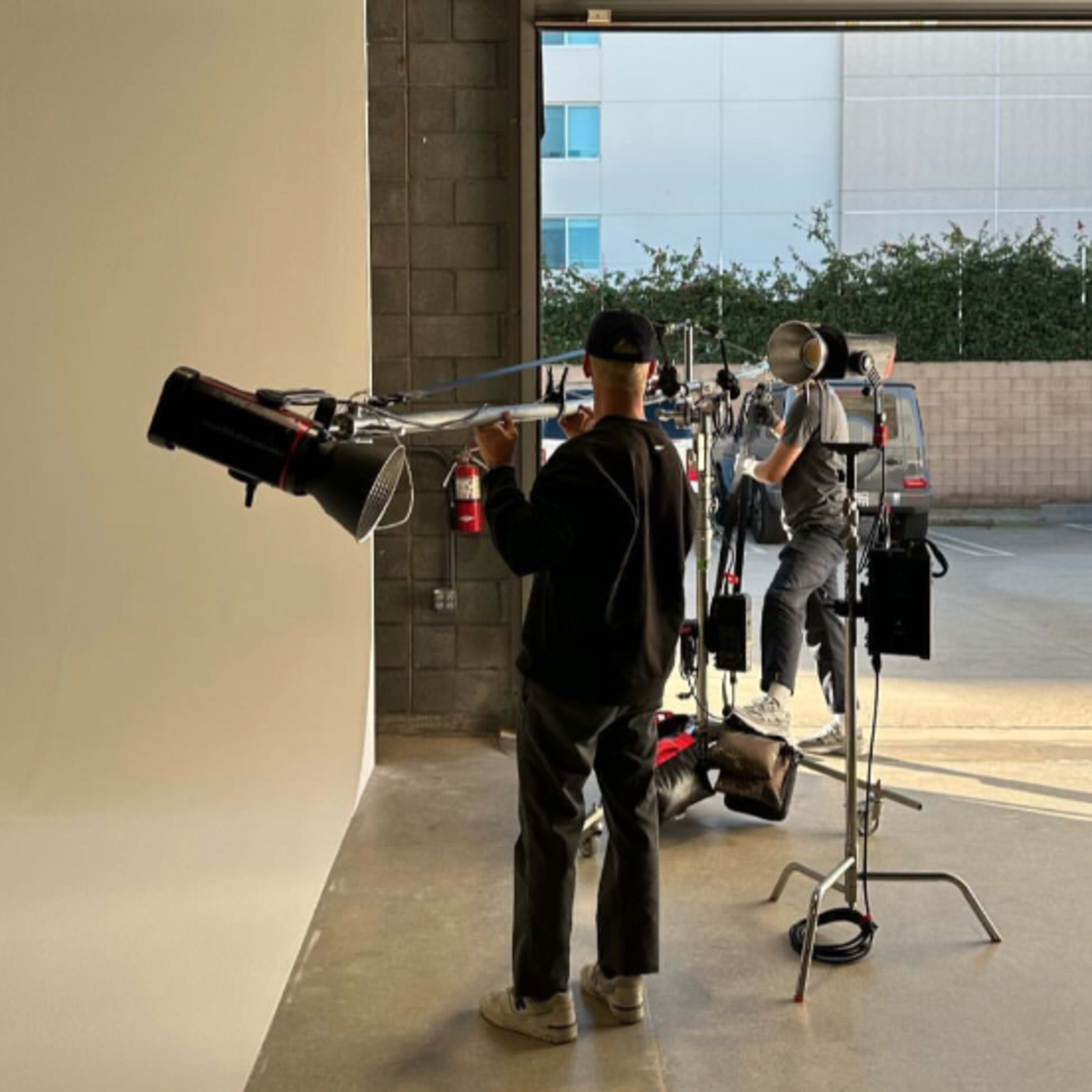 A person operates a camera rig while another adjusts equipment near an open garage door in a studio.