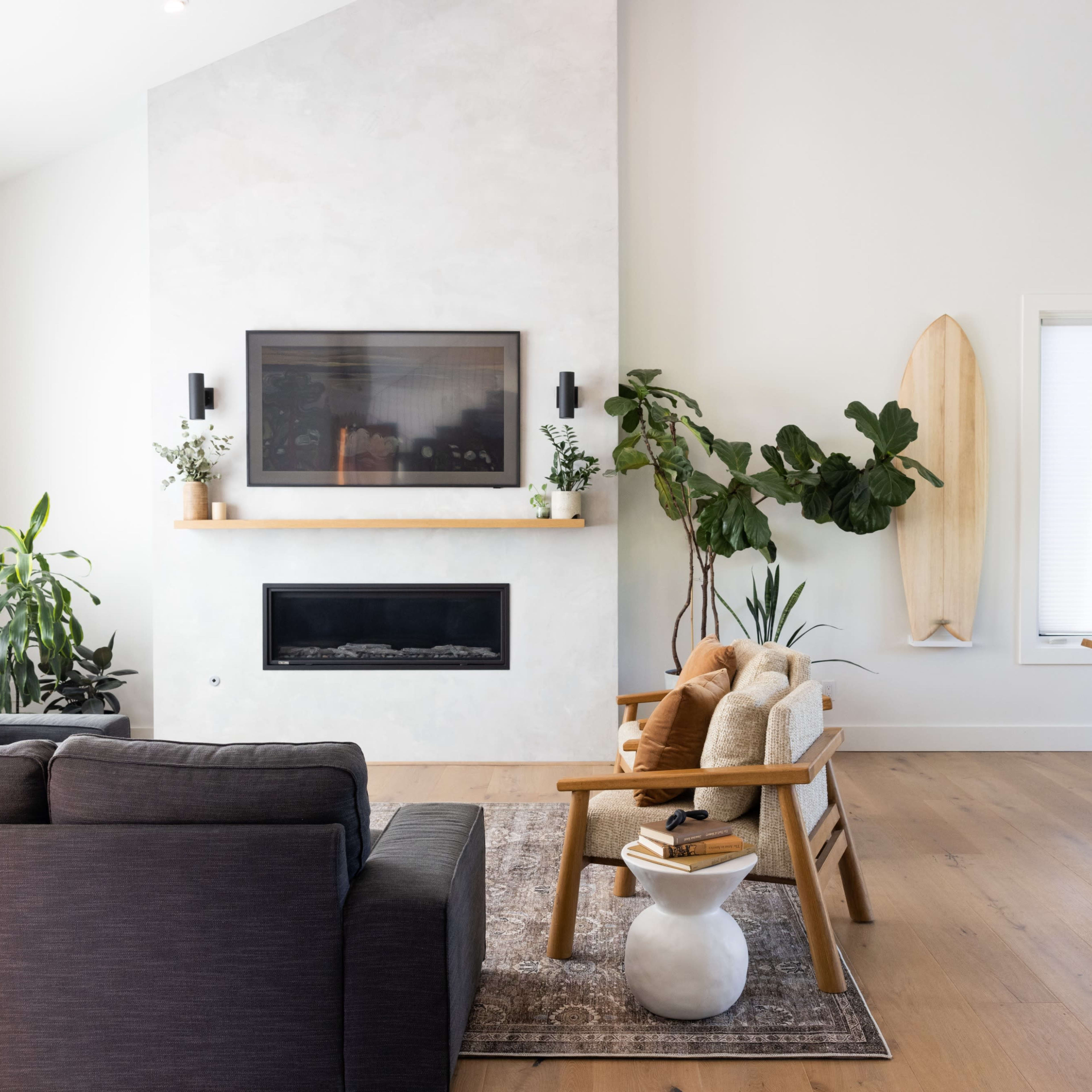 A modern living room featuring a sectional sofa, a coffee table with stacked books, and a dining area with wooden chairs, accompanied by plants and a surfboard on the wall.