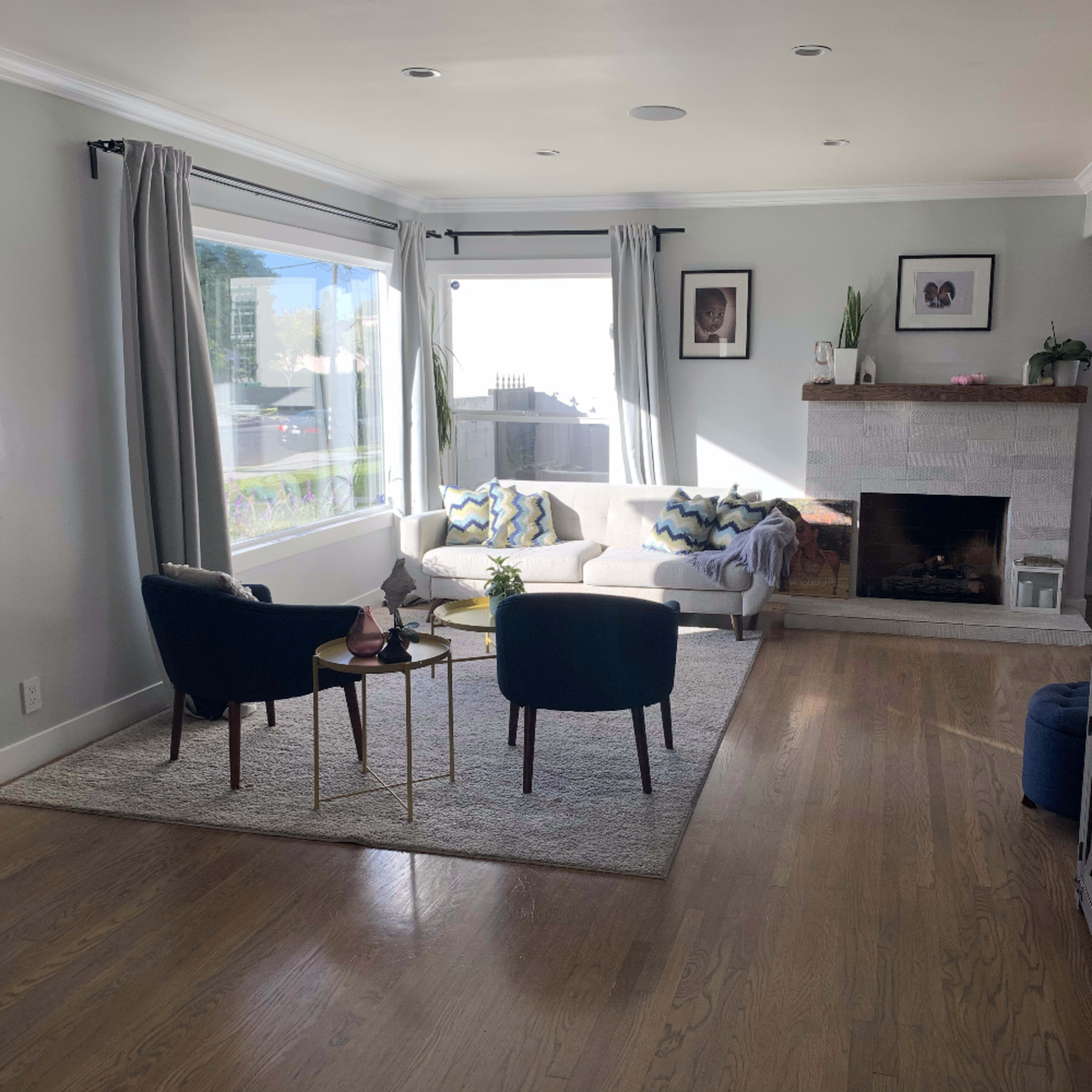 A living room features a large window with sheer curtains, a light-colored sofa, two chairs, a small coffee table, and a stone fireplace.
