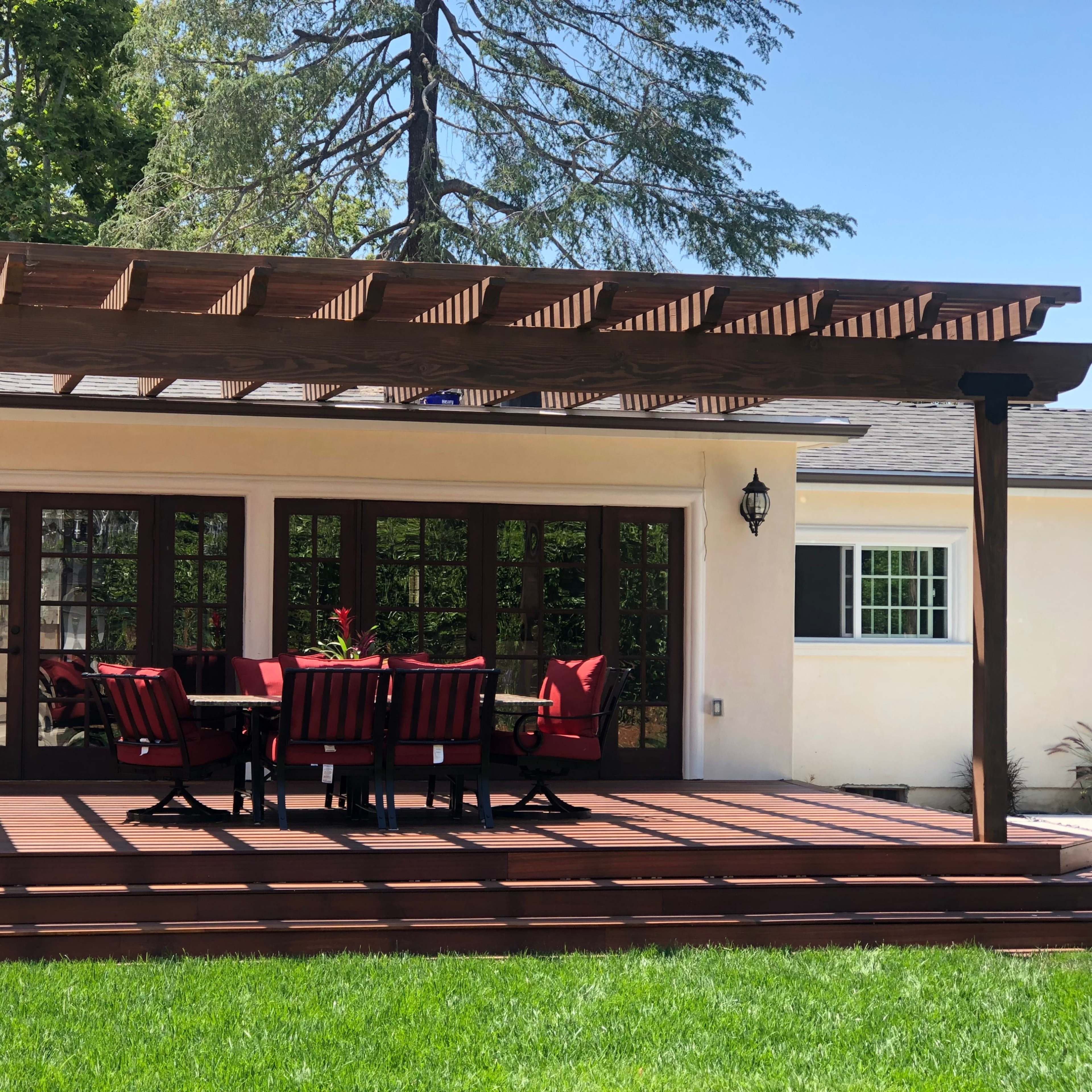 A wooden pergola stands over a dining table with red chairs on a deck next to a house surrounded by grass and trees.