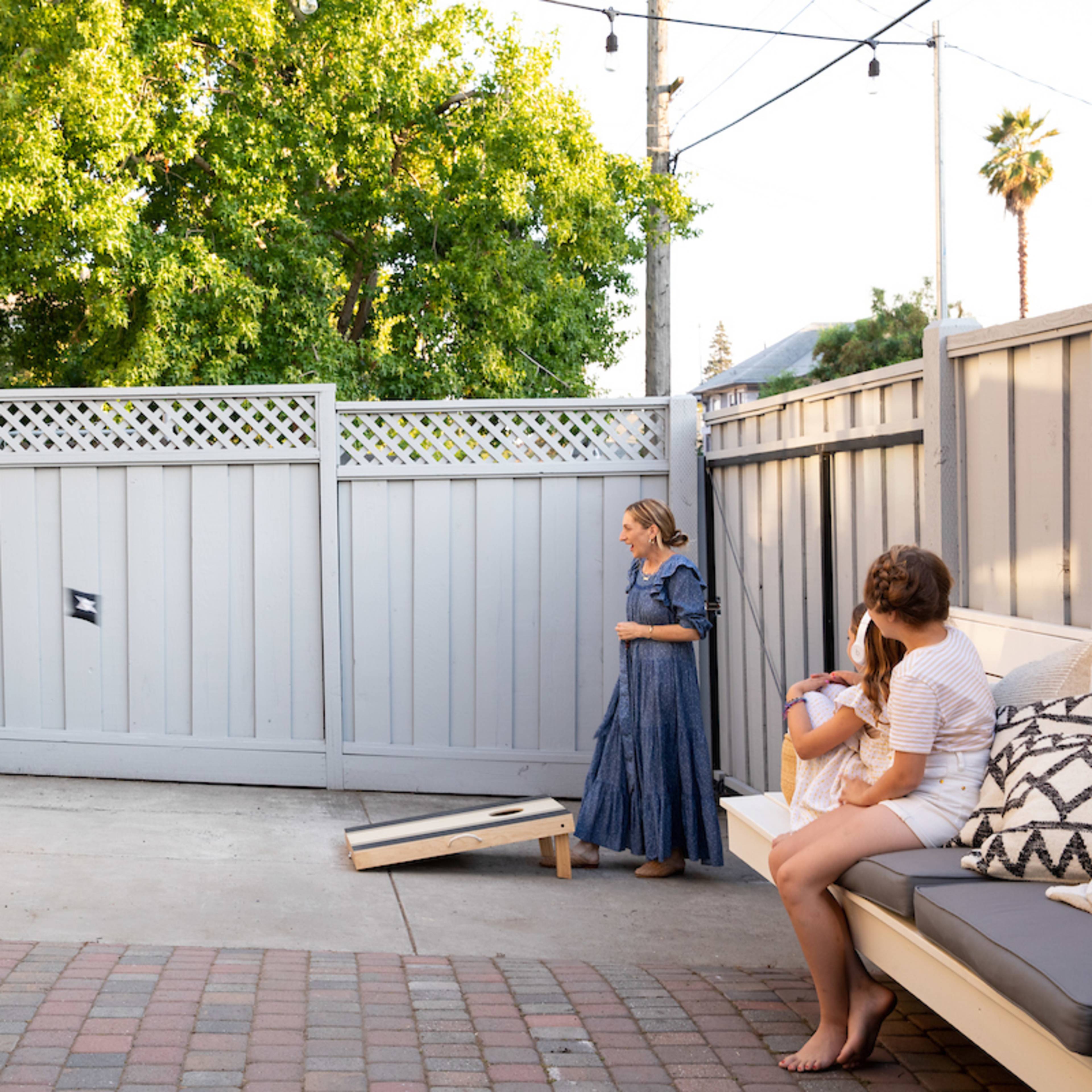 Two people play cornhole in a backyard while a third person sits on a couch nearby.