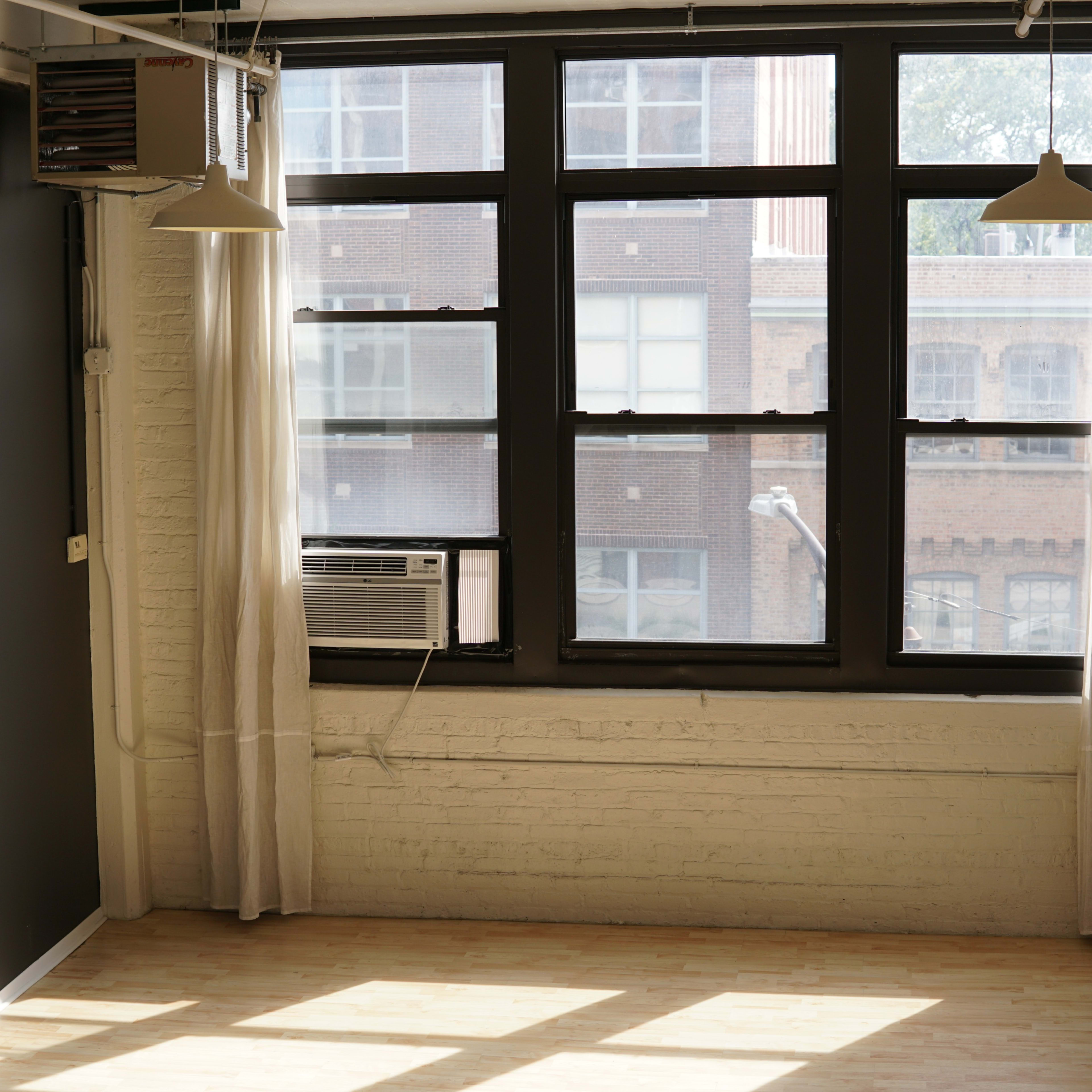 A sunlit, empty room with large windows featuring an air conditioning unit and light curtains.