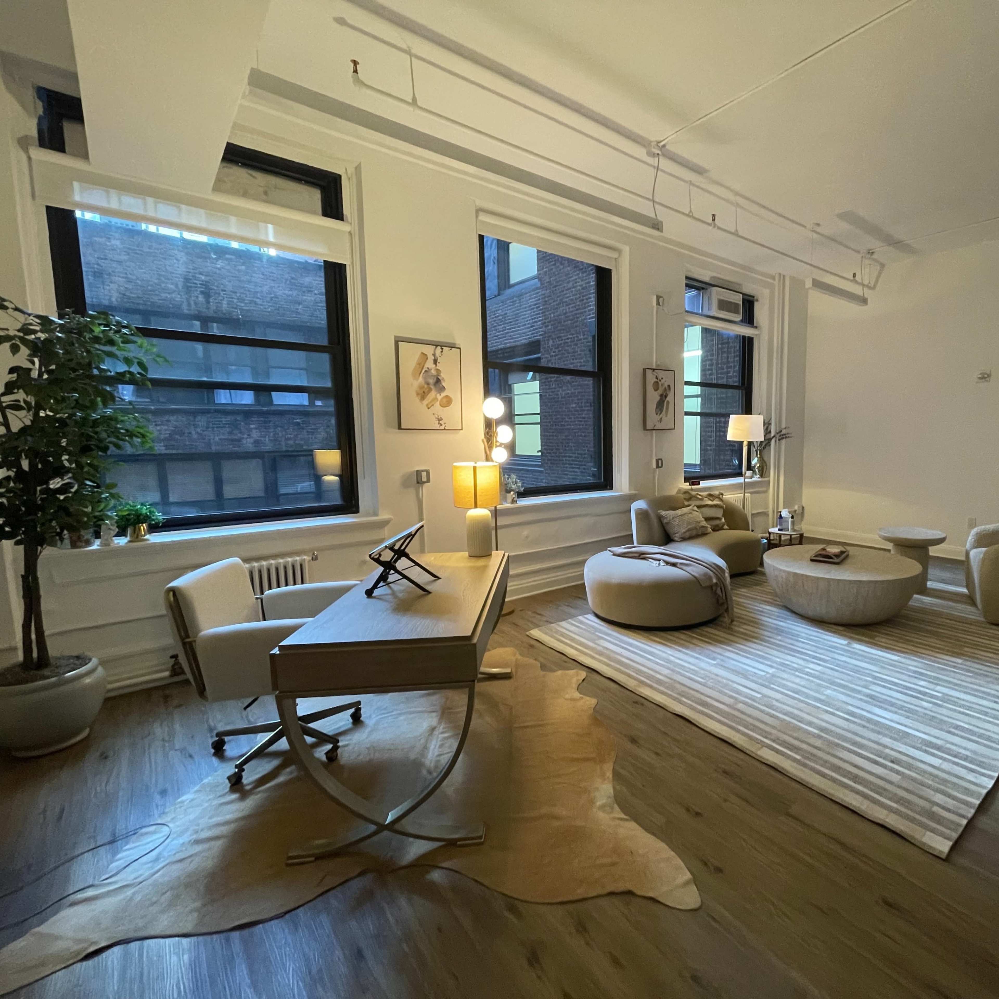 A modern office space with a desk, a seating area featuring round poufs and armchairs, large windows, and a decorative plant.