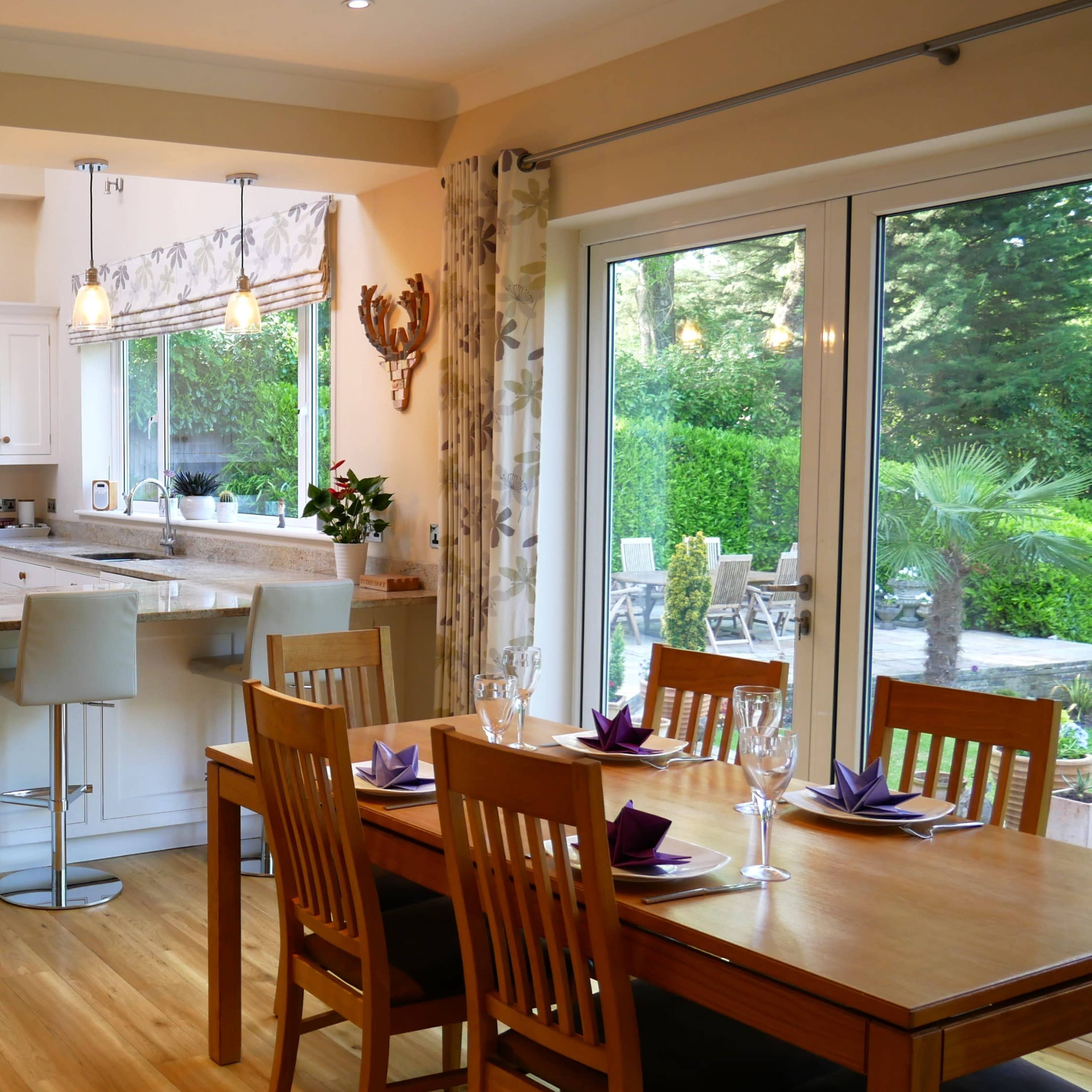 A spacious kitchen and dining area feature wooden flooring, modern appliances, and a dining table set for a meal beside large glass doors leading to a garden.