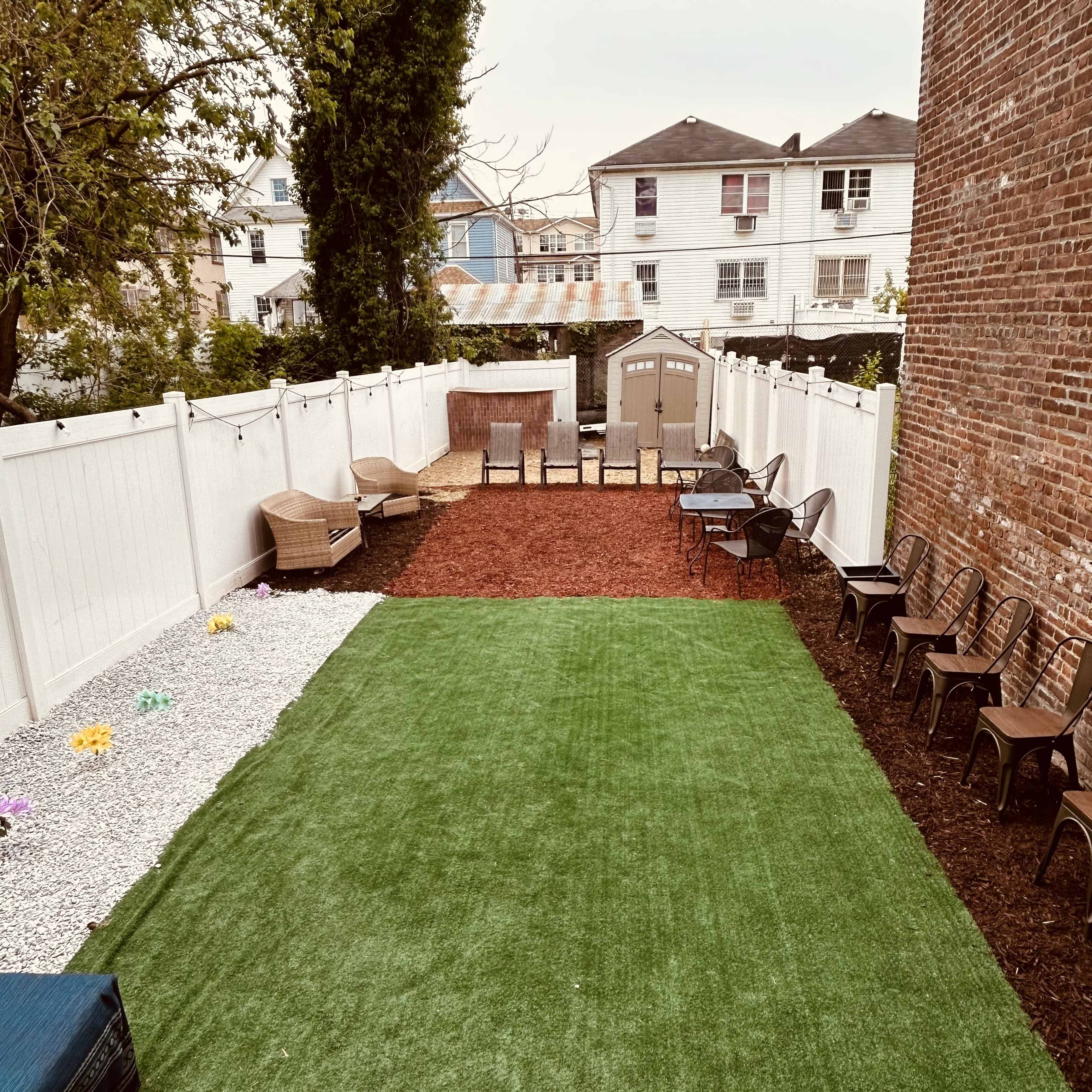 A backyard with a section of artificial grass, bordered by gravel and mulch, featuring several chairs and a storage shed in the background.