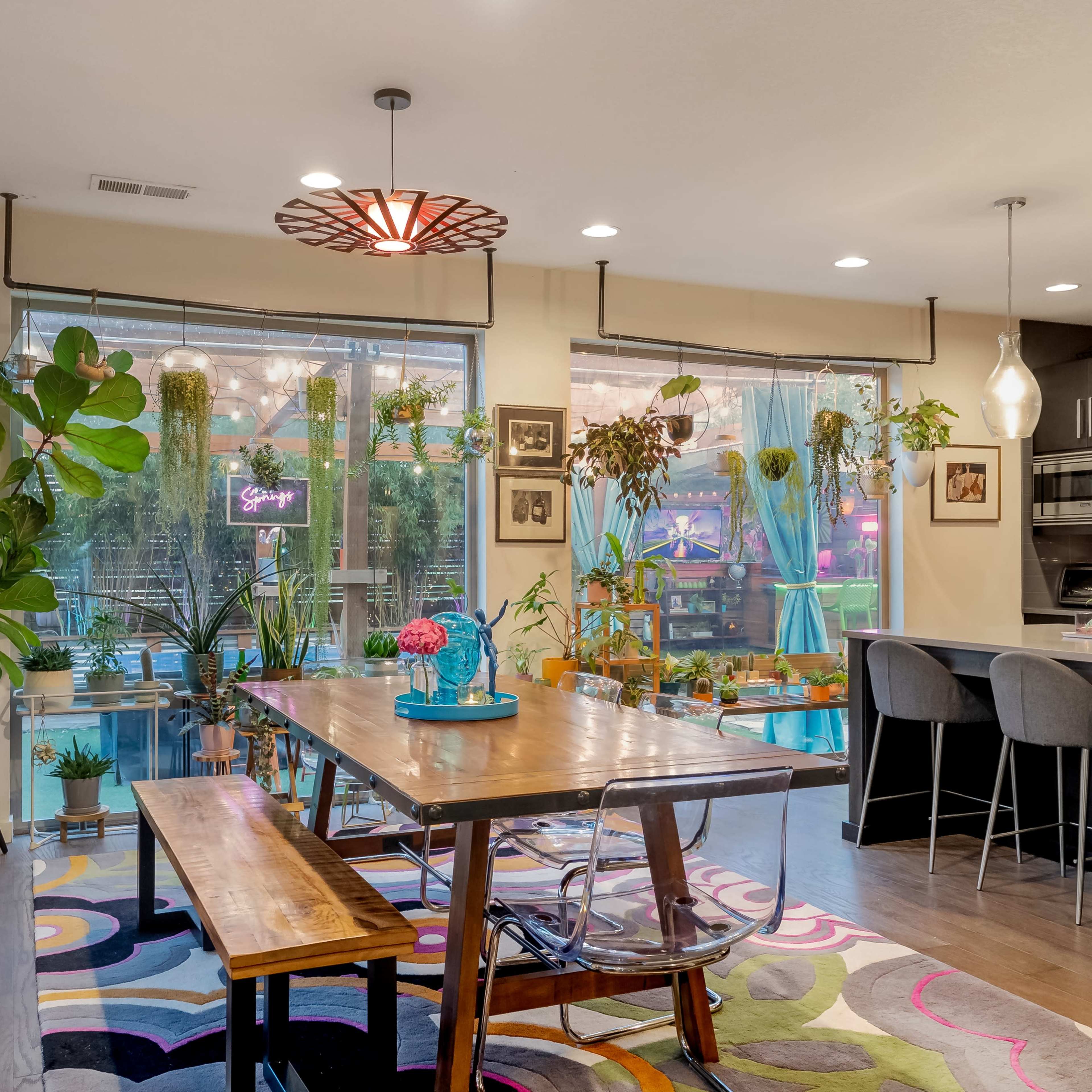A modern dining area featuring a large wooden table surrounded by clear chairs, with green plants and colorful decor elements throughout the space.