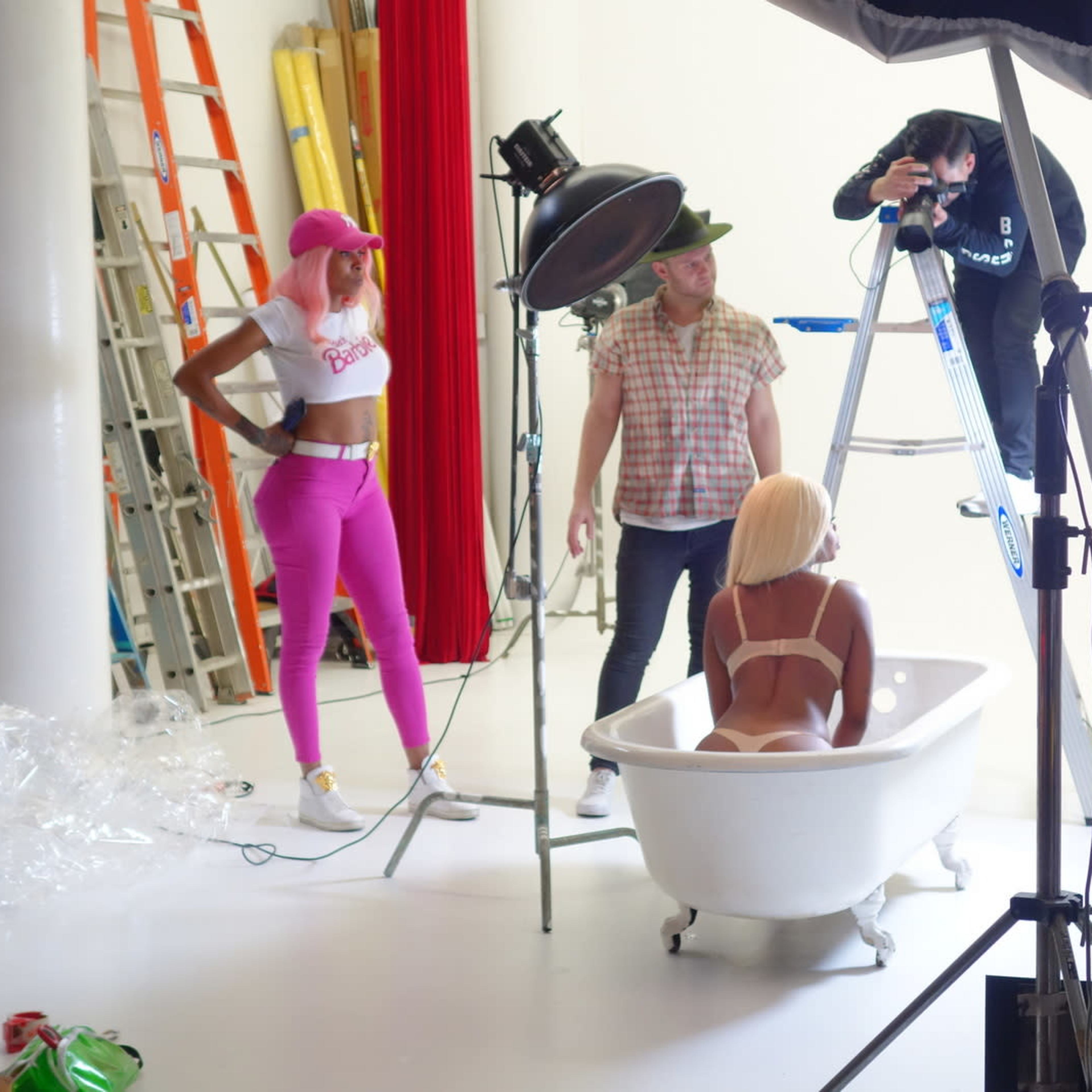 A photoshoot is taking place in a studio with a model in a bathtub, while a photographer sets up equipment and two other individuals assist.