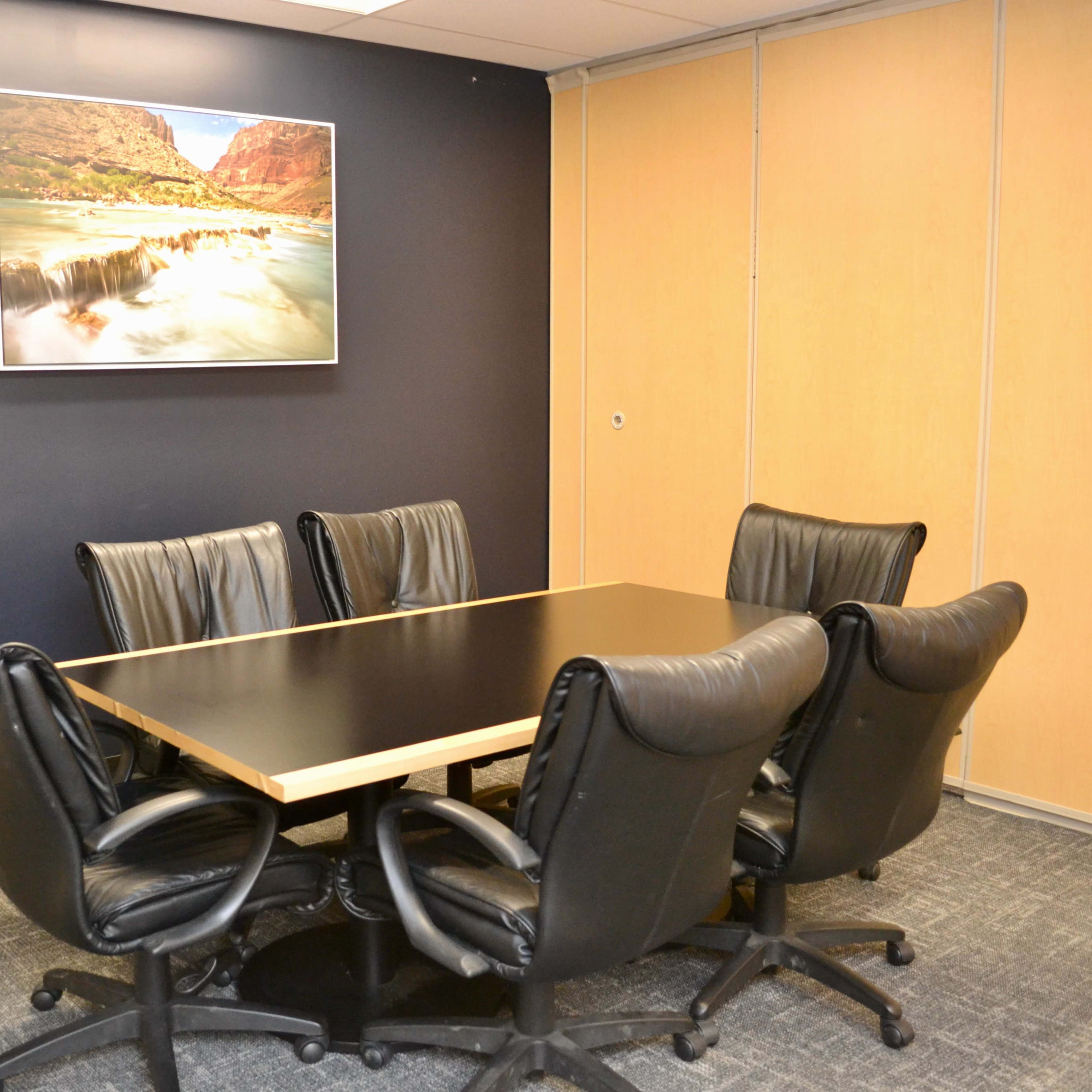 A conference room with a large black table surrounded by six leather office chairs, and a landscape picture hung on the wall above.