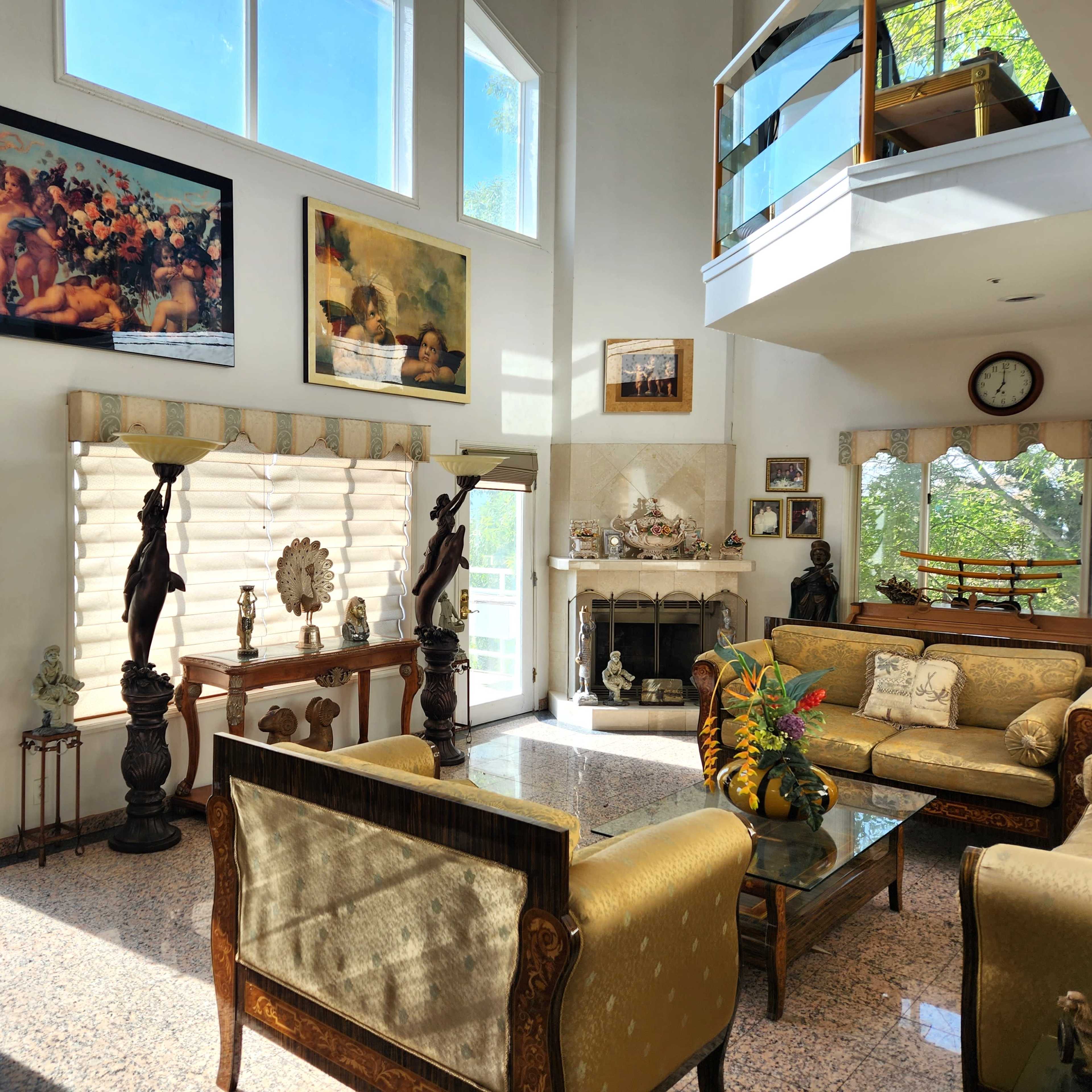 The living room features a high ceiling, large windows, and a mix of decorative art, with seating arrangements centered around a coffee table.