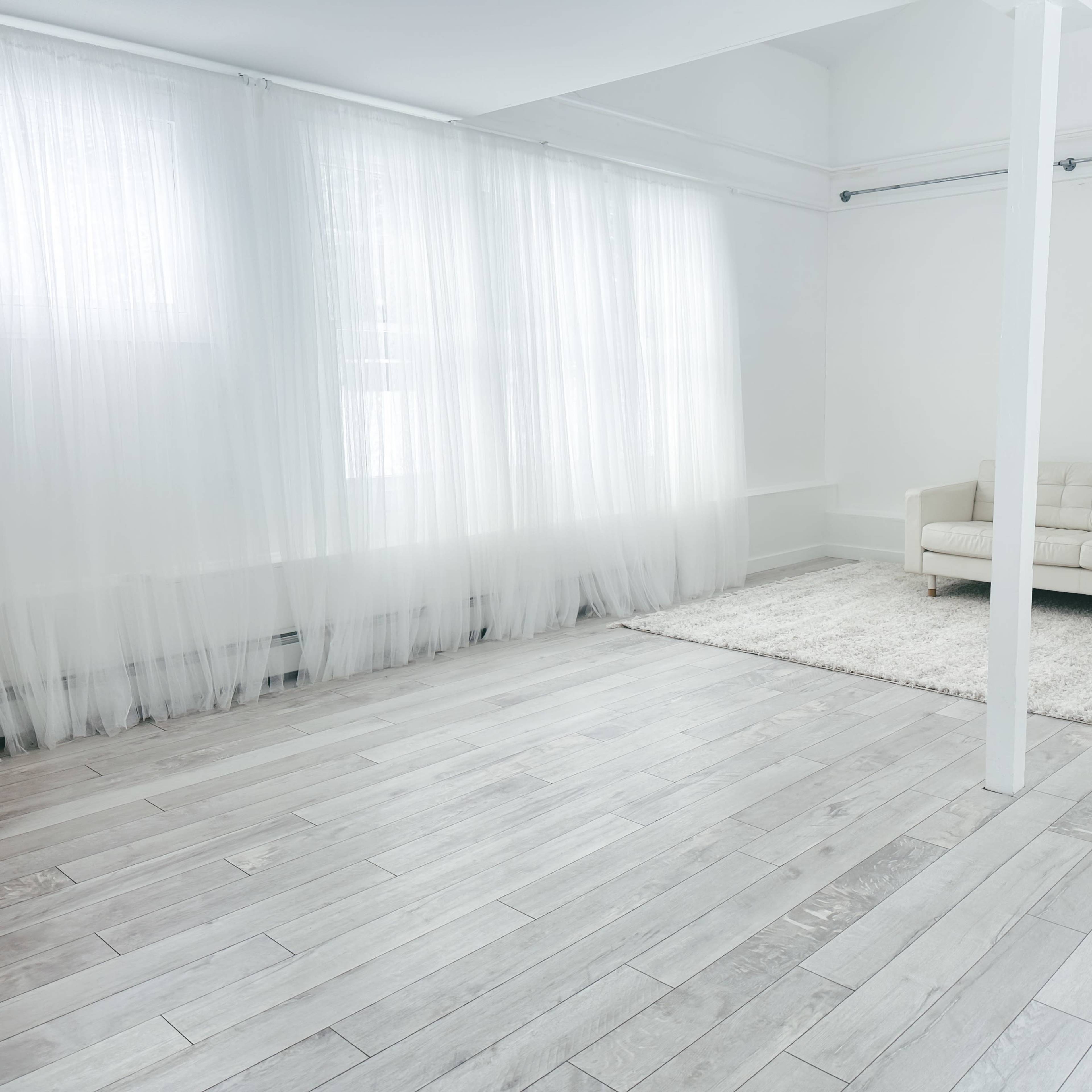 A bright, minimalist room with white walls, light-colored wooden flooring, a plush area rug, and a modern sofa near sheer curtains.