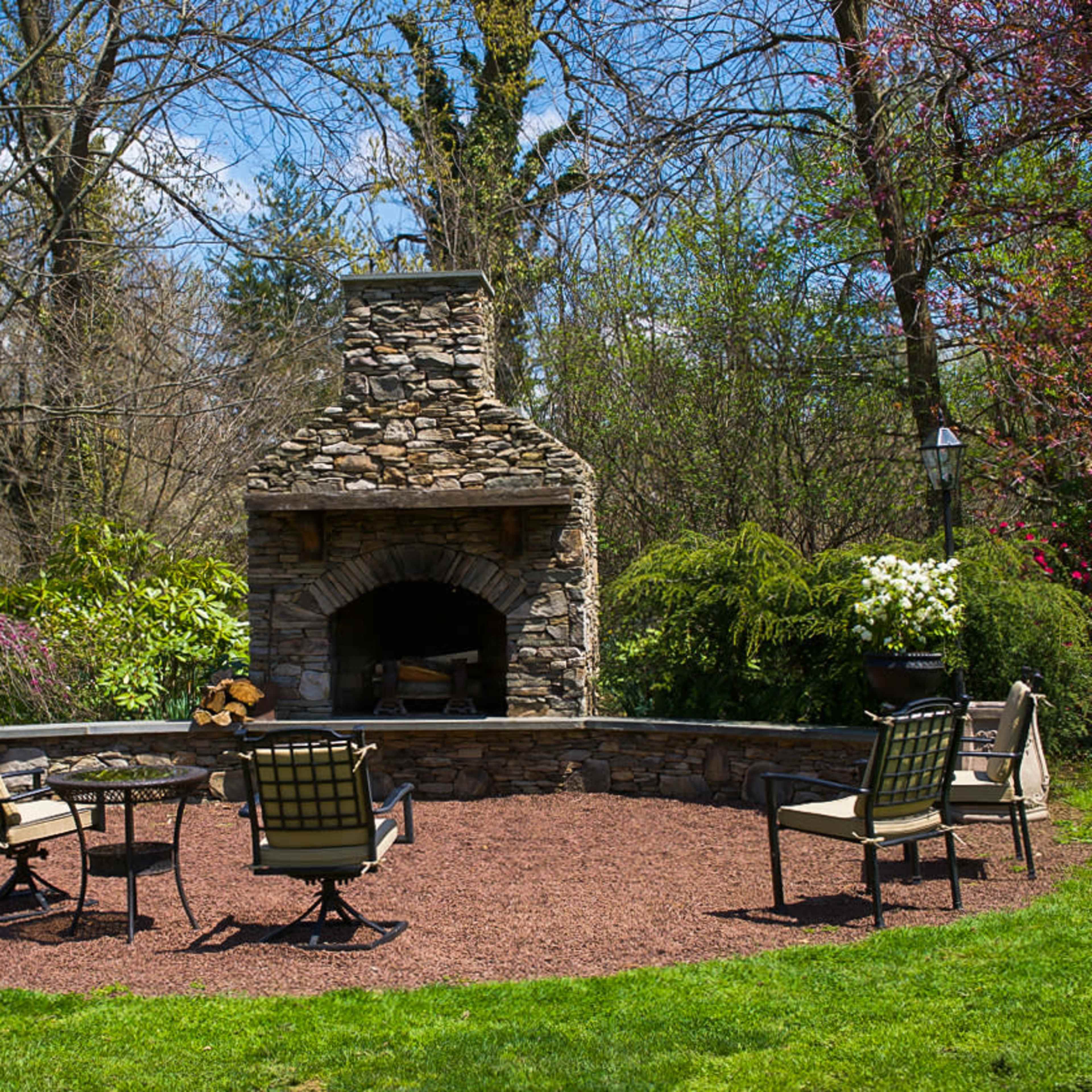 A stone outdoor fireplace is surrounded by several chairs on a gravel patio, with trees and flowering plants in the background.