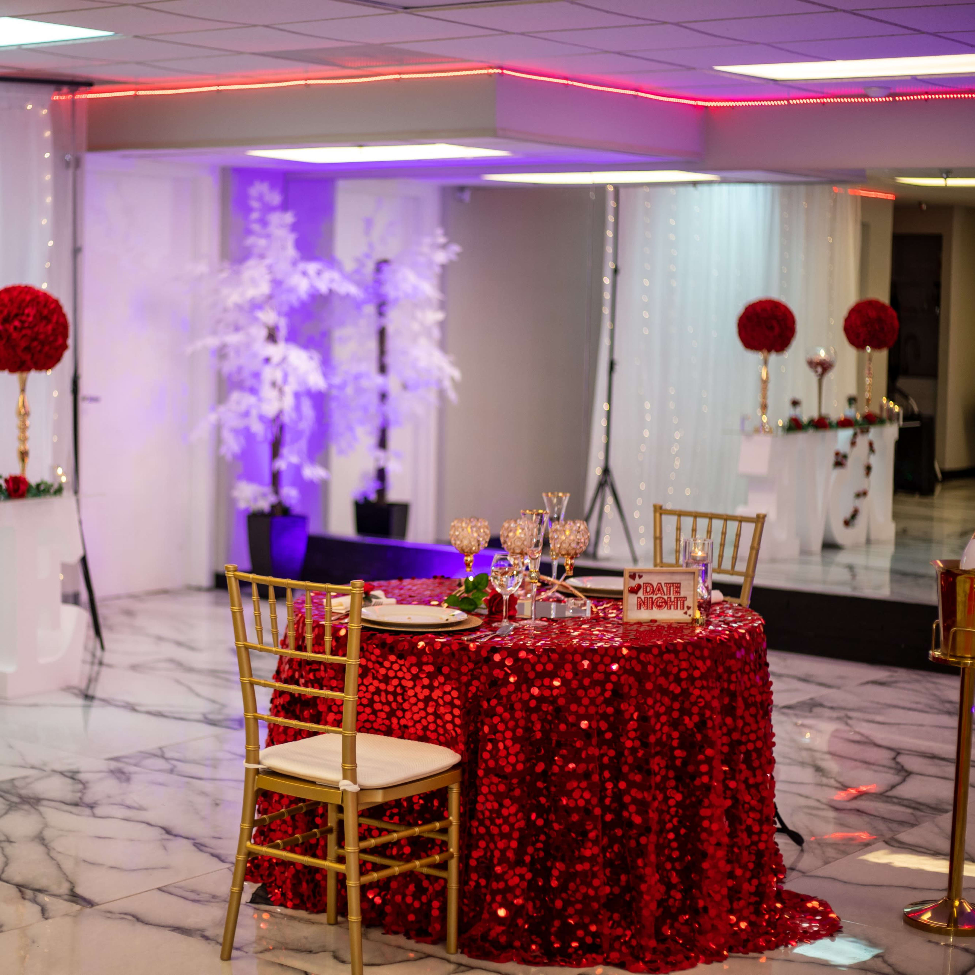 A decorated event space featuring a table covered in red sequins, surrounded by gold chairs, with floral arrangements and a large "LOVE" sign in the background.