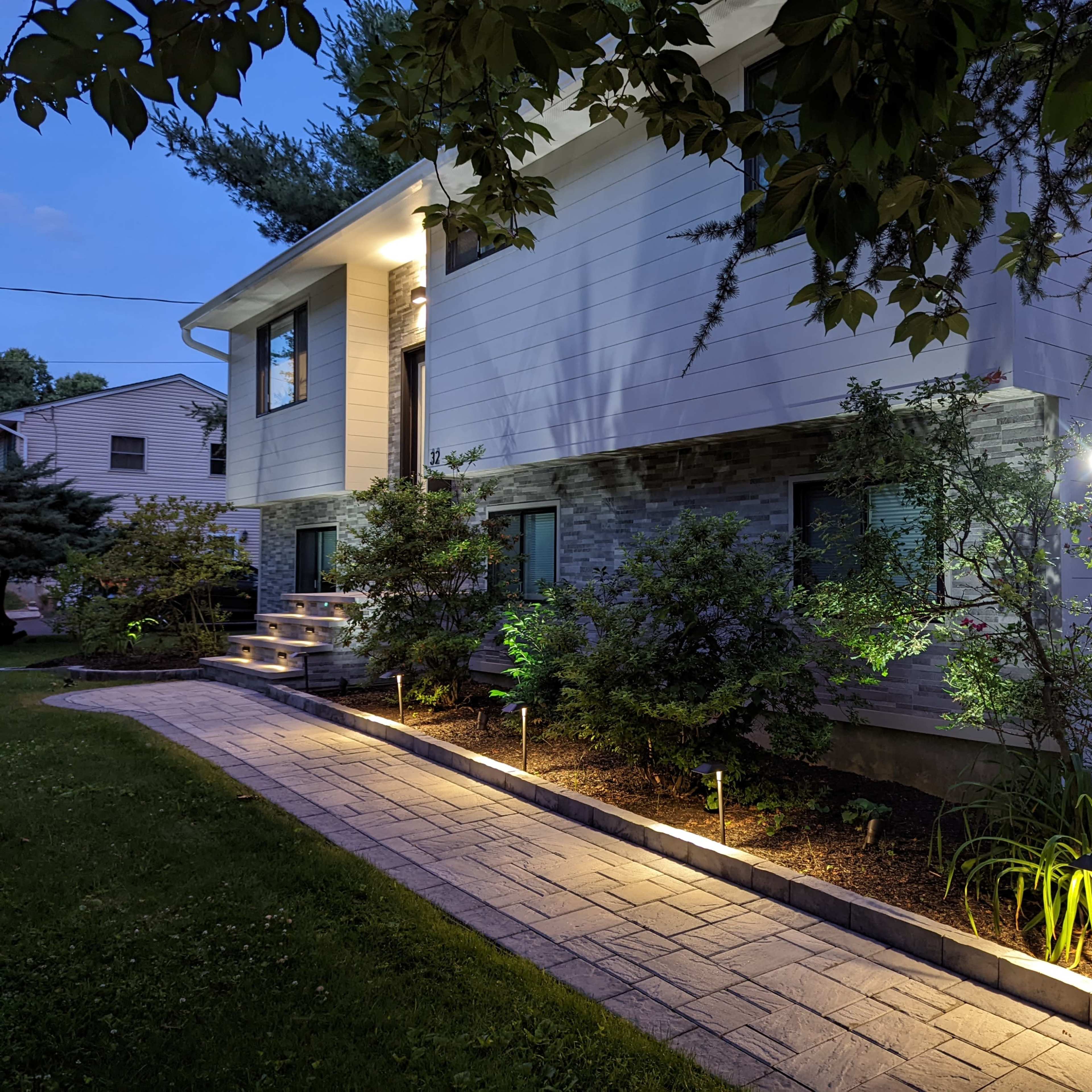 A well-lit pathway leads to a modern house with a landscaped front yard in the evening.