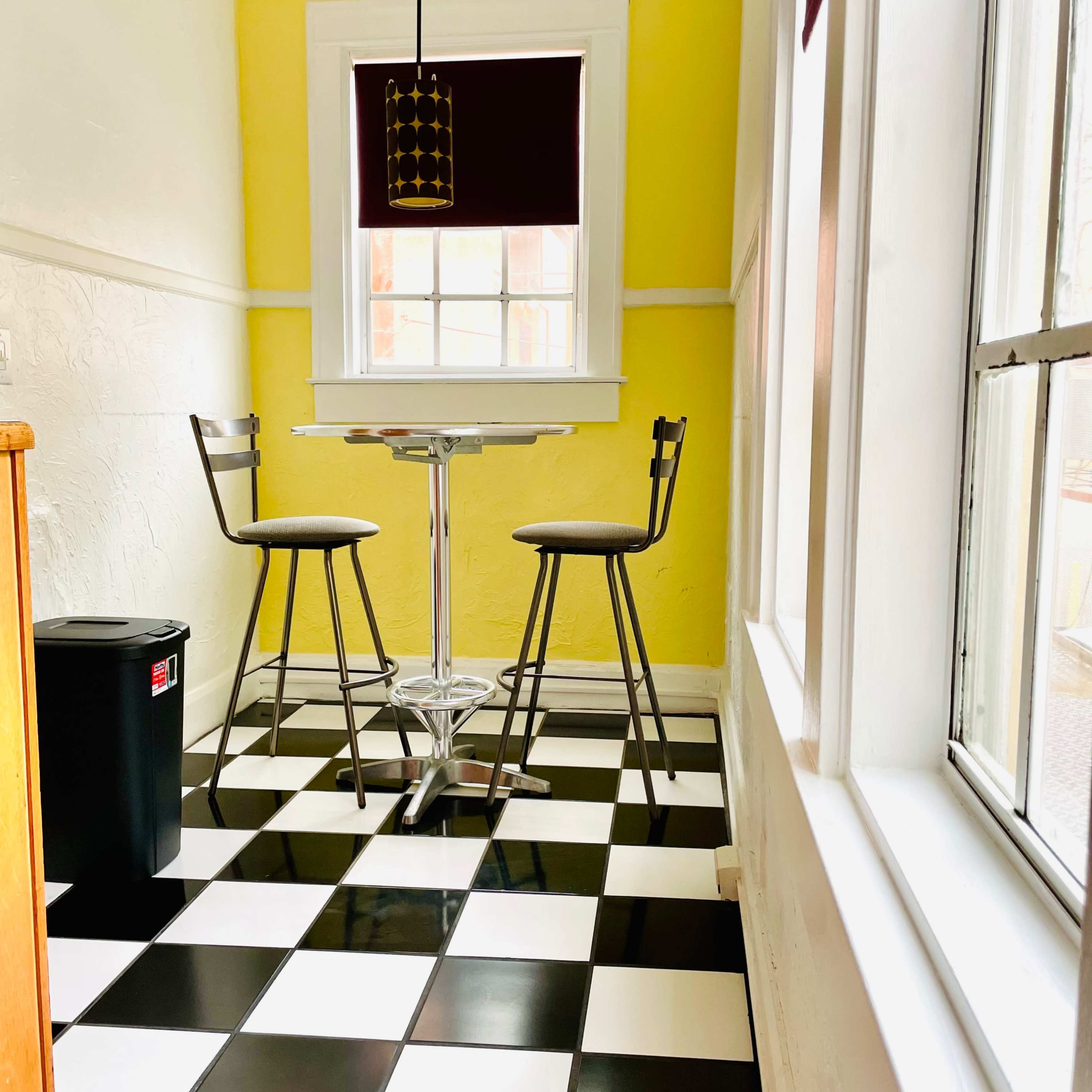 A small kitchen nook features a round table and two high stools against a bright yellow wall, with black and white checkered flooring.