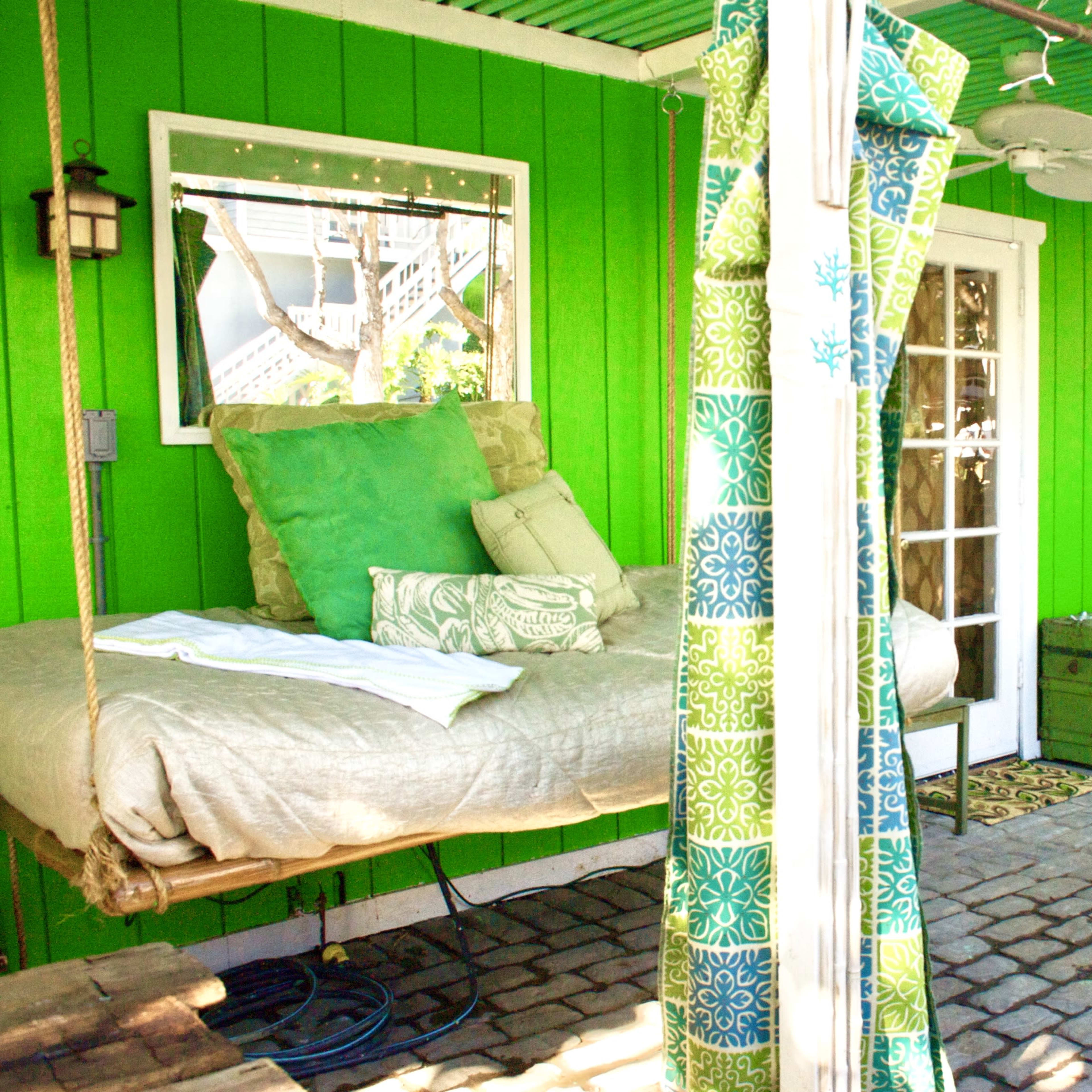 A vibrant green outdoor area with a swing bed, a round chair, and patterned curtains.