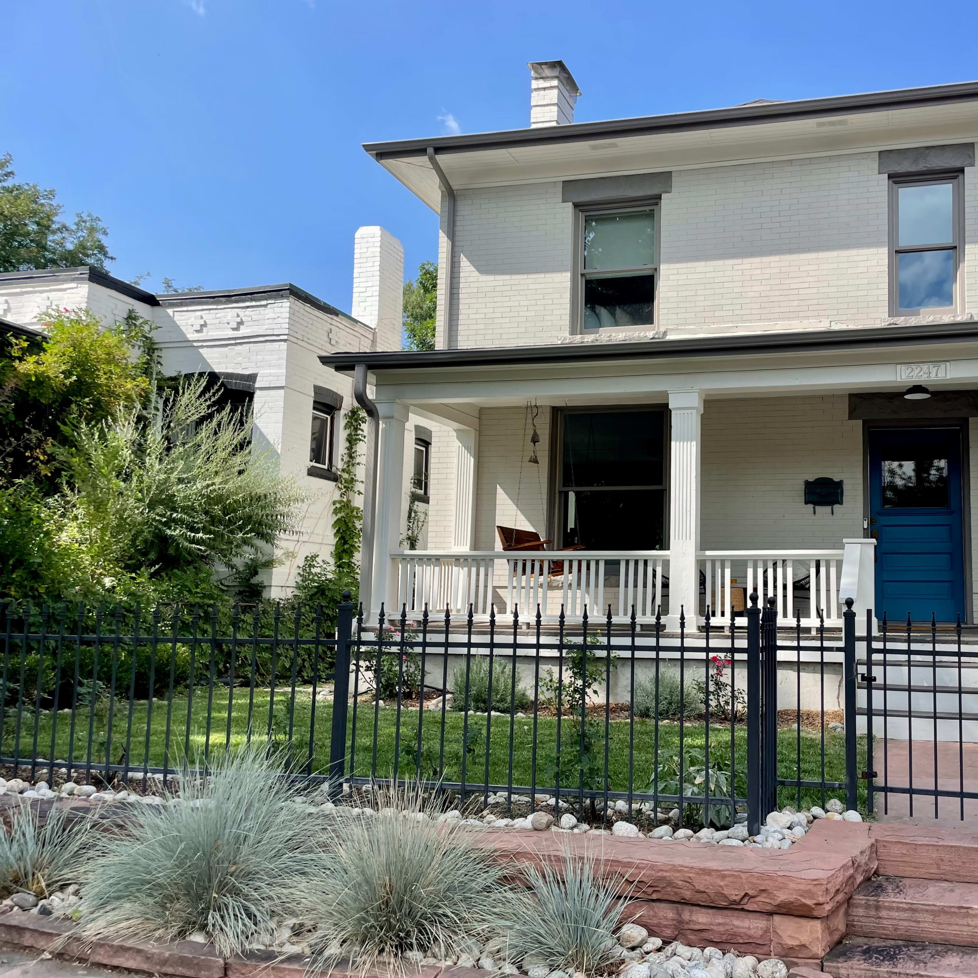 A two-story white house with a blue front door and a porch, surrounded by a manicured garden and a black wrought-iron fence.