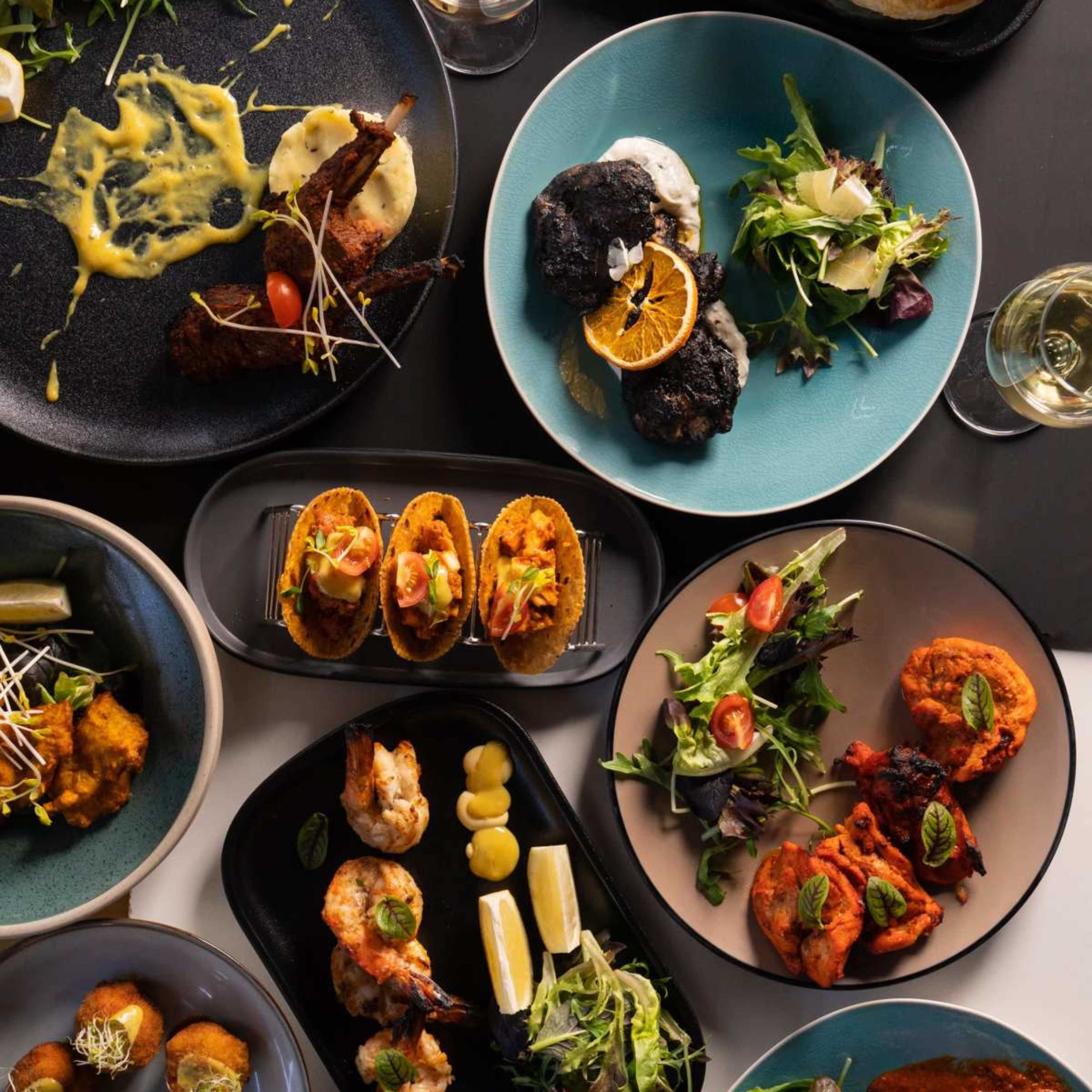 A variety of dishes arranged on a table, including tacos, grilled shrimp, salads, and a pastry, alongside drinks and sauces.