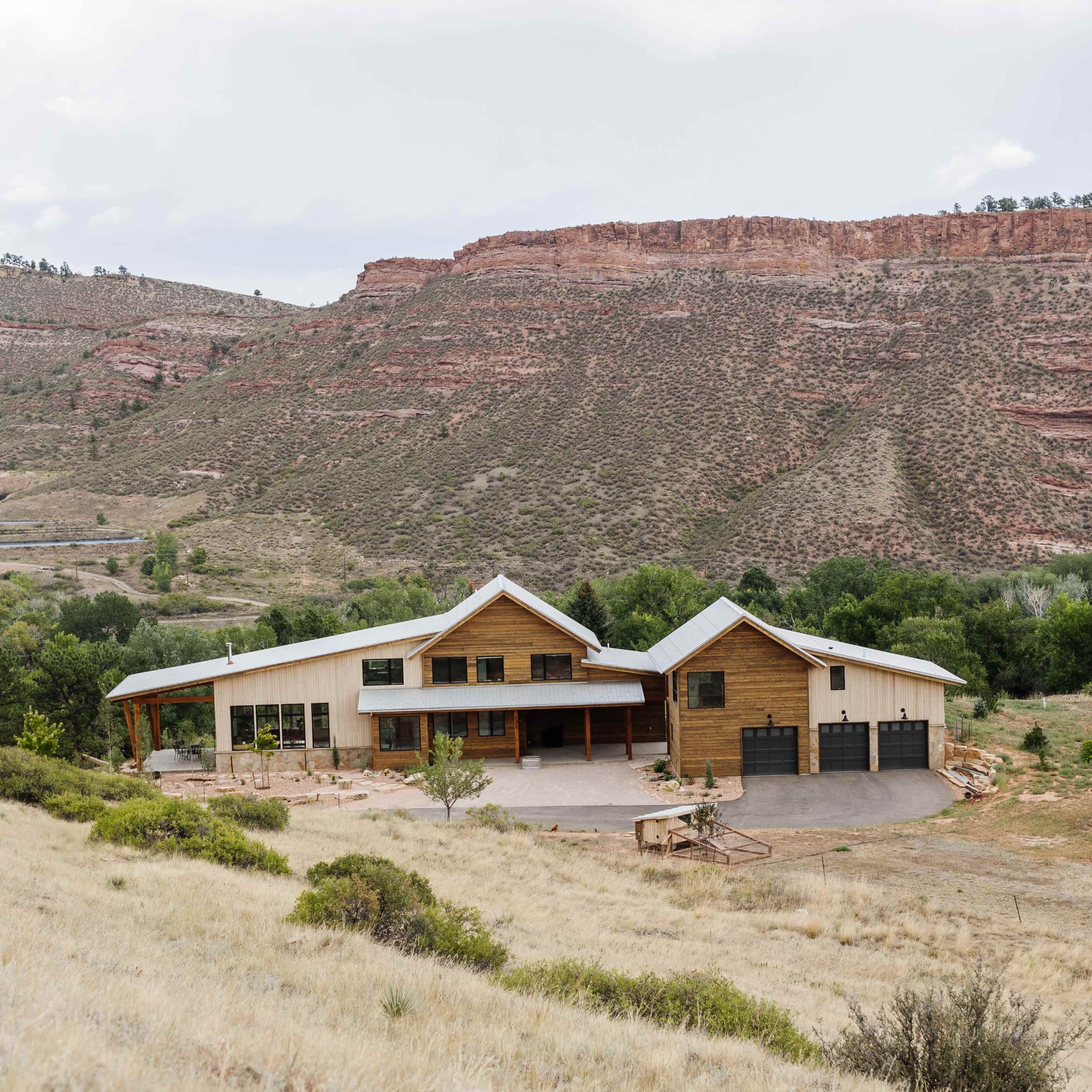 A modern house with a mix of wood and stucco exterior is situated on a hillside, overlooking a valley with rocky outcrops and sparse vegetation.