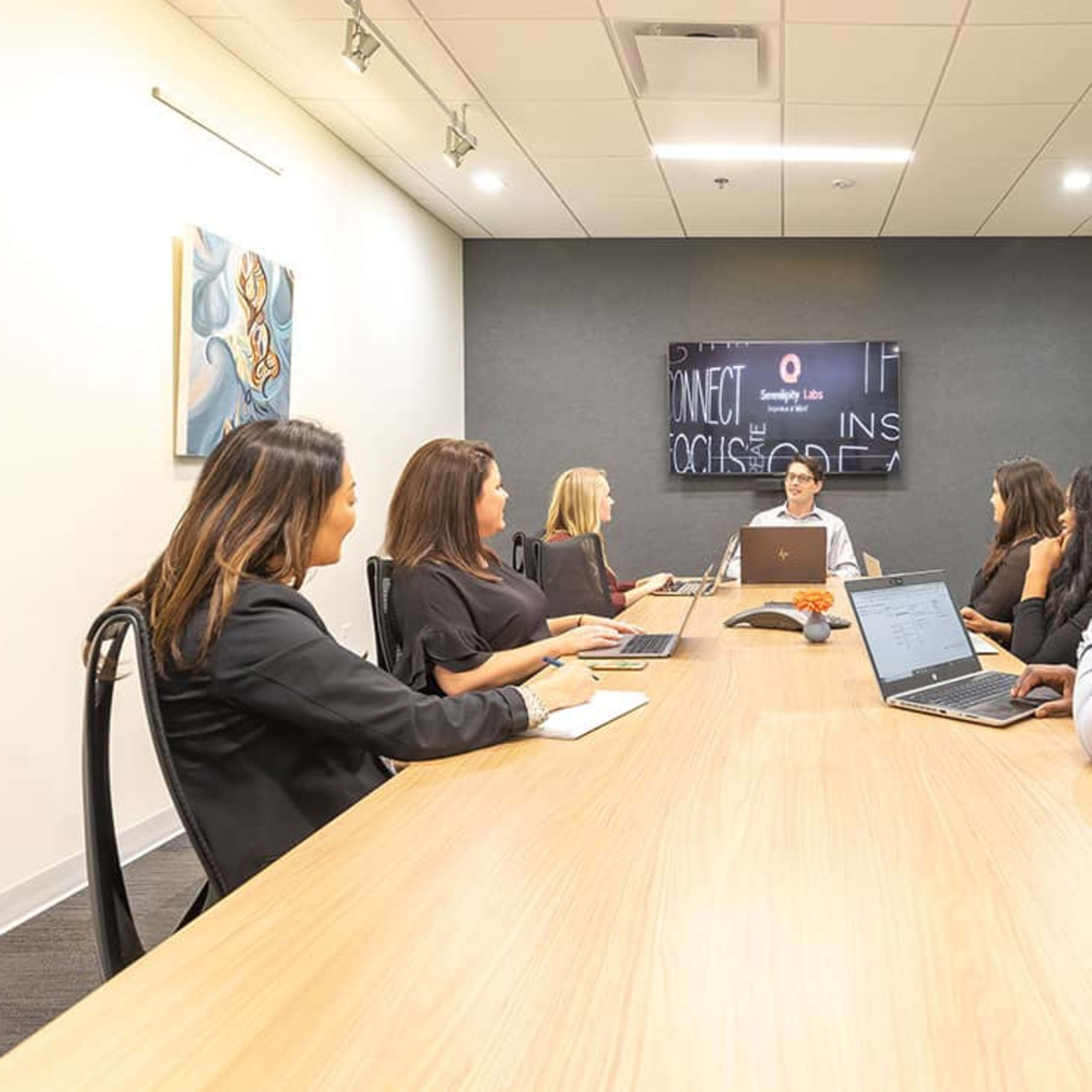A group of six professionals sit around a long conference table in a modern meeting room, with a screen displaying information at the front.