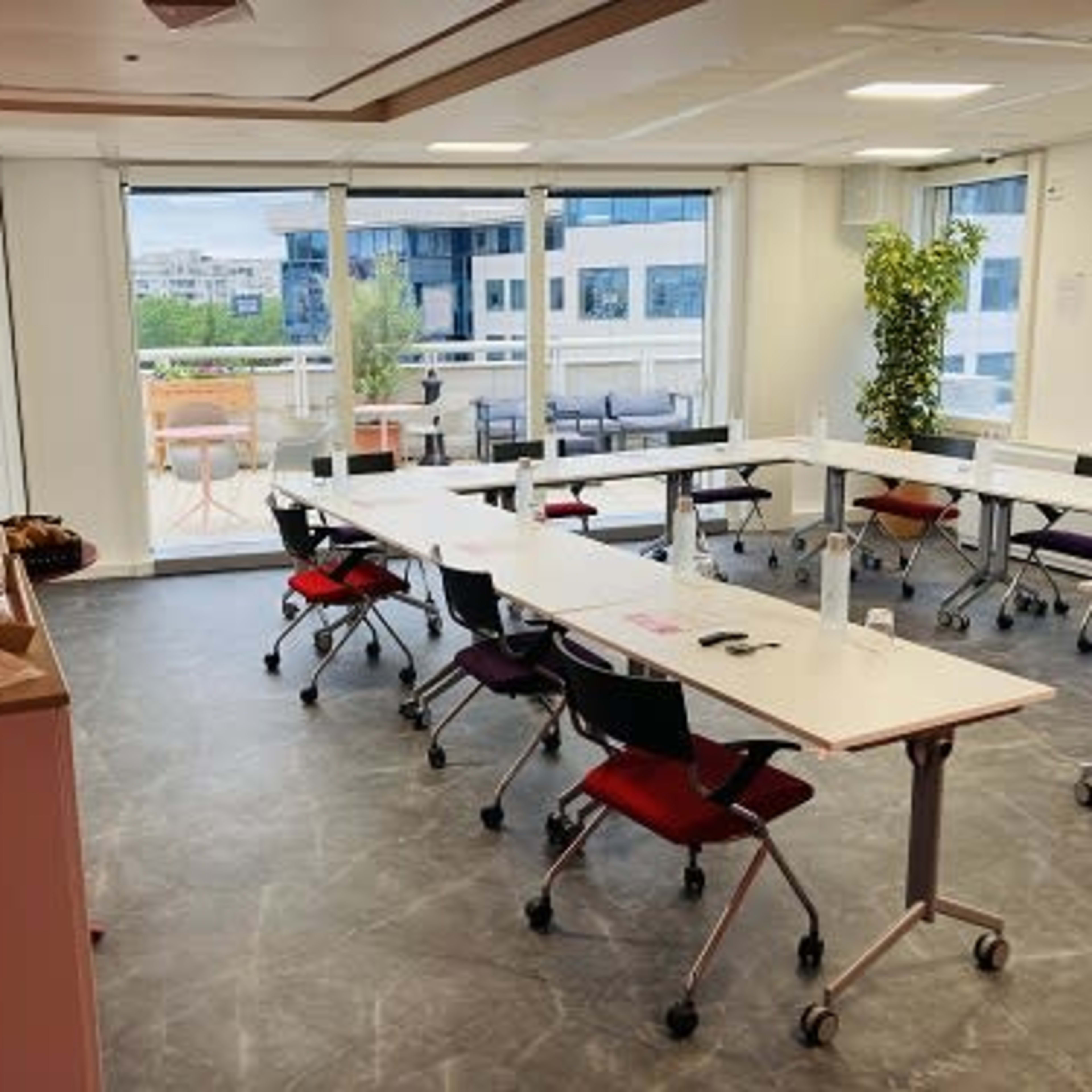 A modern meeting room with several tables arranged for a conference, featuring large windows that provide a view of the outside space with greenery.