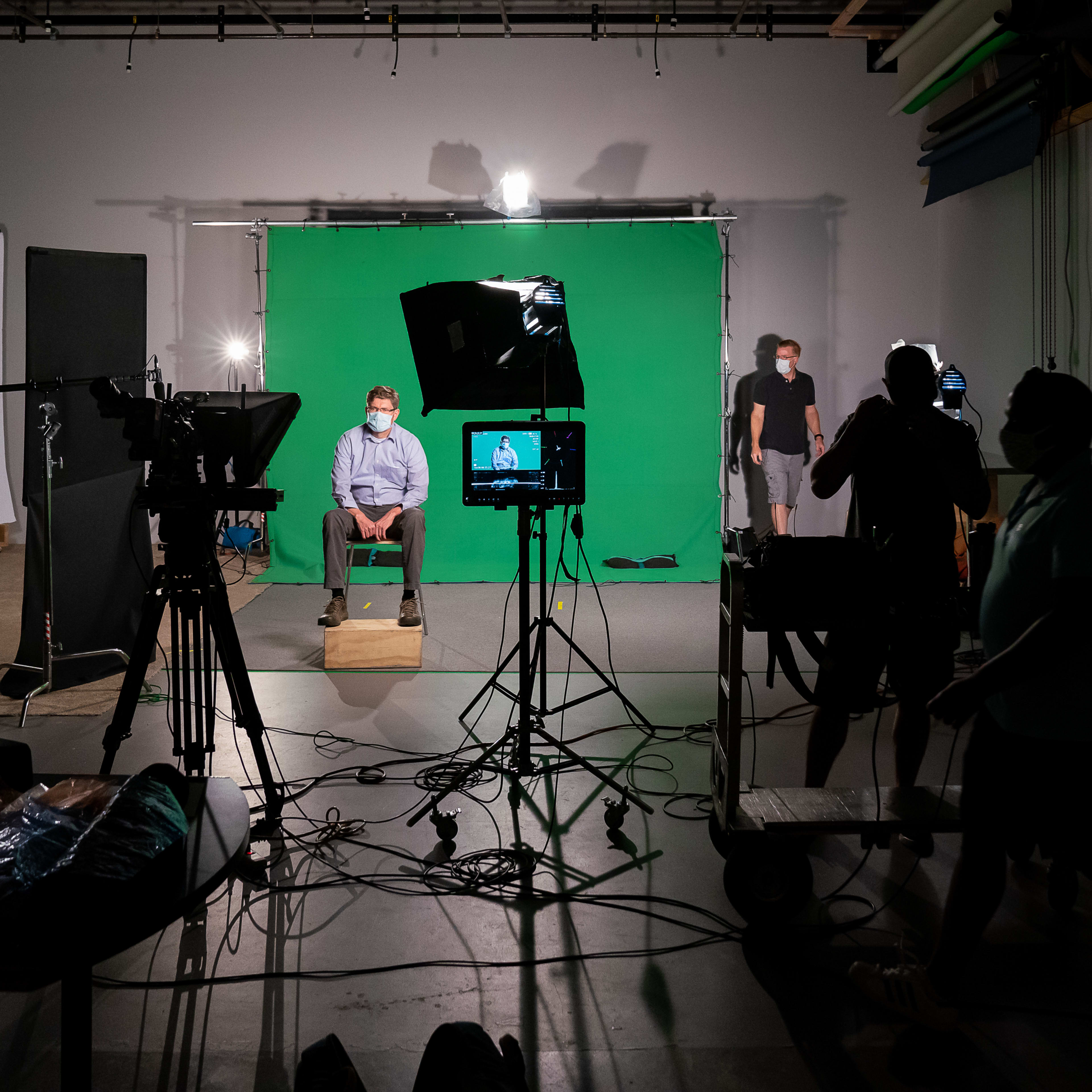 A person sits on a wooden box in front of a green screen while multiple camera setups and crew members are positioned nearby.